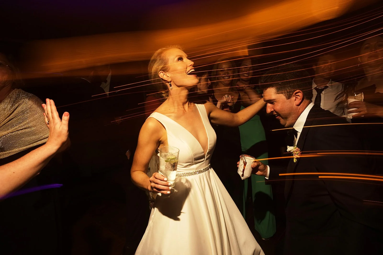 A woman in a white wedding dress smiling and holding a drink, dancing at a wedding reception with a man in a tuxedo, surrounded by celebrating guests.