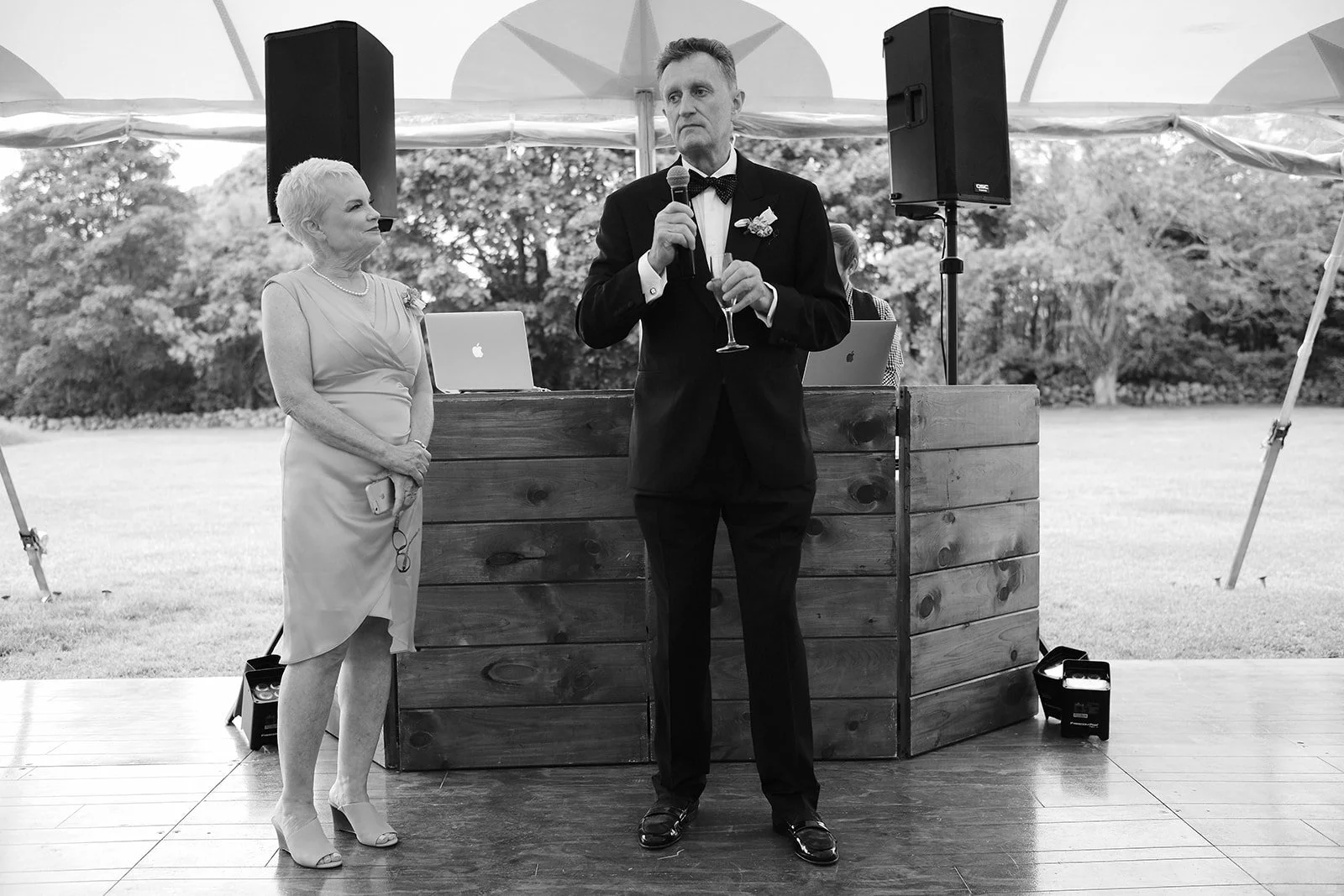 A man giving a speech at a wedding reception outdoors under a canopy, with a woman standing beside him. The man is in a tuxedo holding a microphone and a glass of champagne. The woman is dressed in a formal dress, holding glasses, and listening atten