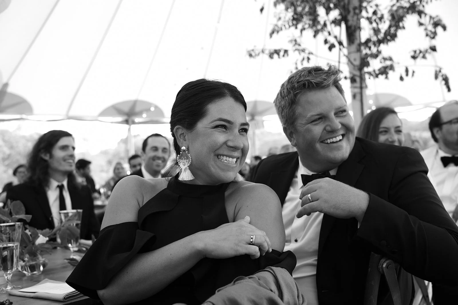 People at a formal event, smiling and laughing, seated under a tent.