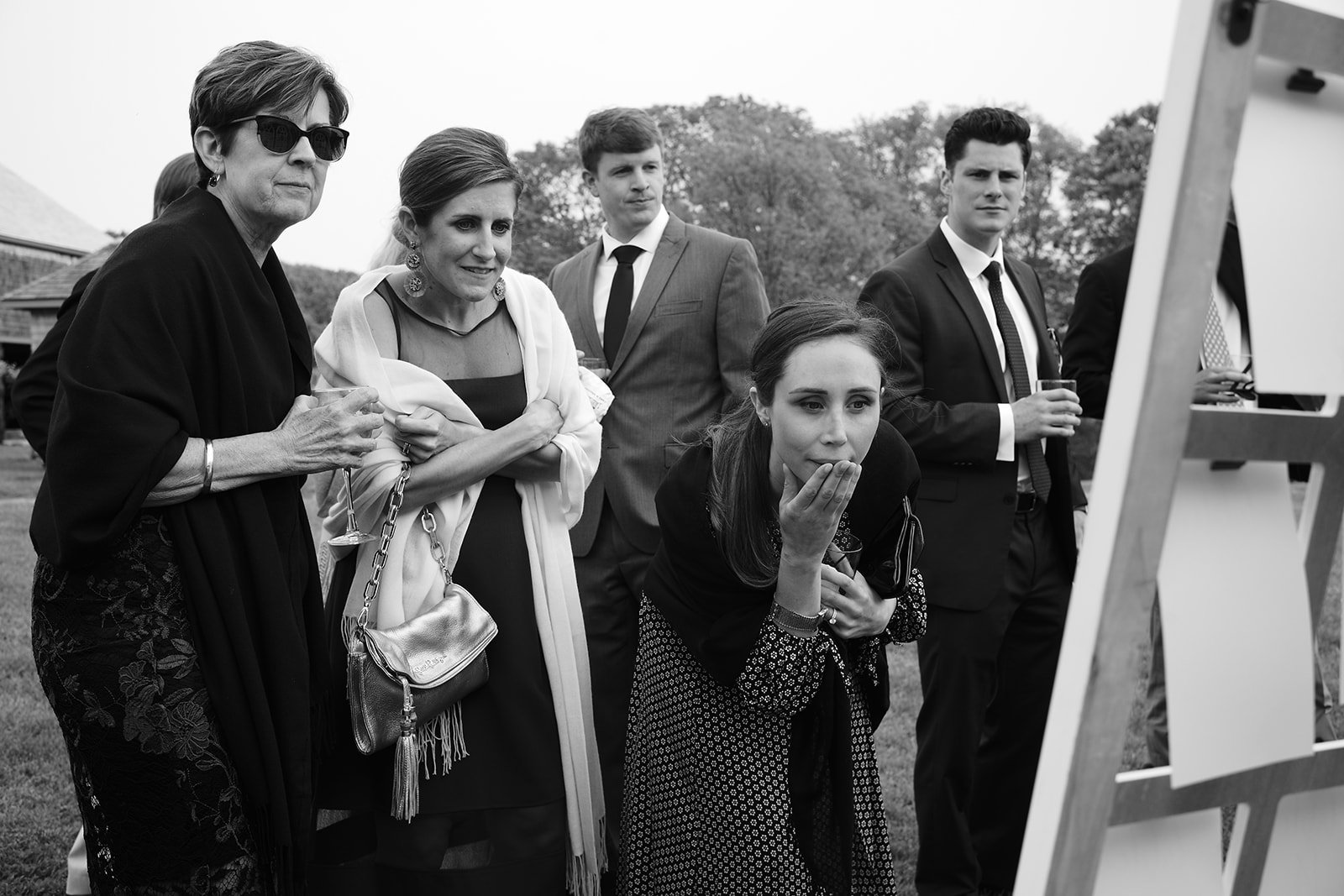 A group of six people, including women and men, dressed in formal attire, gathered outdoors around an easel, observing a presentation or artwork, with trees in the background.