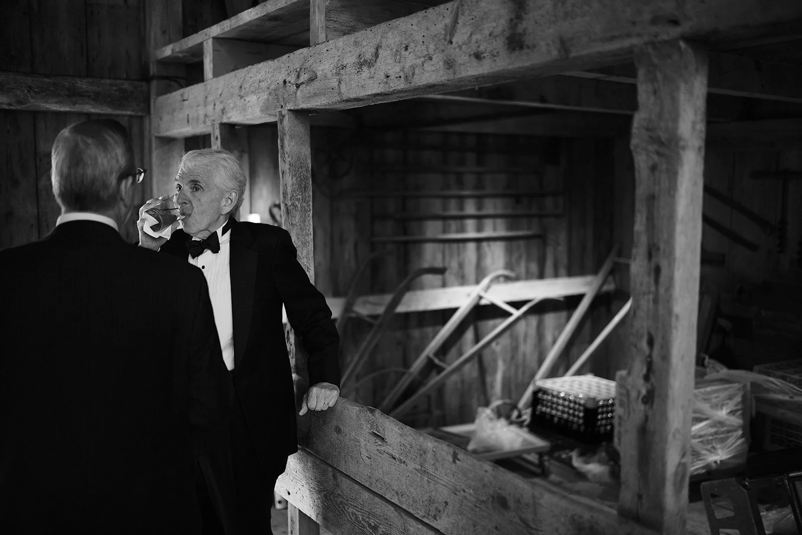 Two men in tuxedos talking inside a barn or rustic shed. One is drinking from a glass.