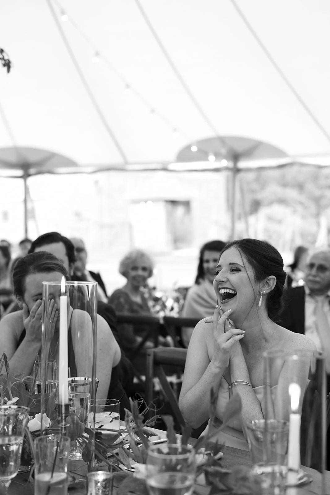 A joyful woman laughing at a wedding reception with guests seated around her, decorated with candles and flowers.