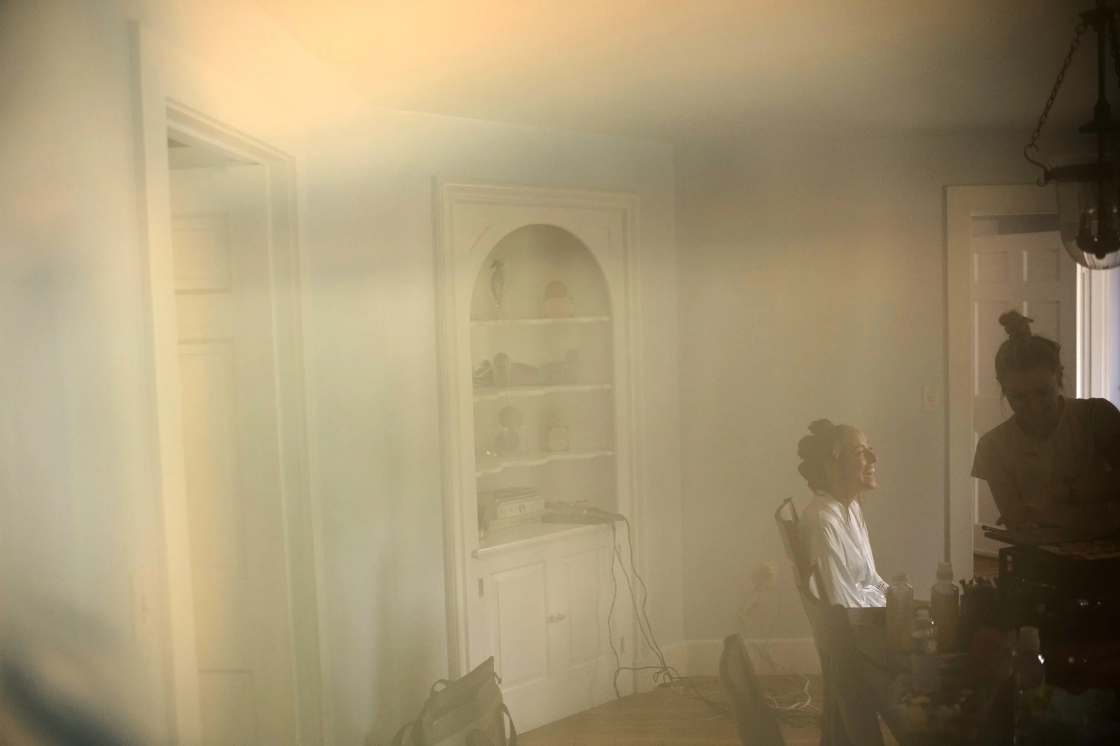 Two women in a room, one seated and smiling, the other standing and looking down, with items on a table and a white built-in cabinet in the background, seen through a foggy or frosted window.