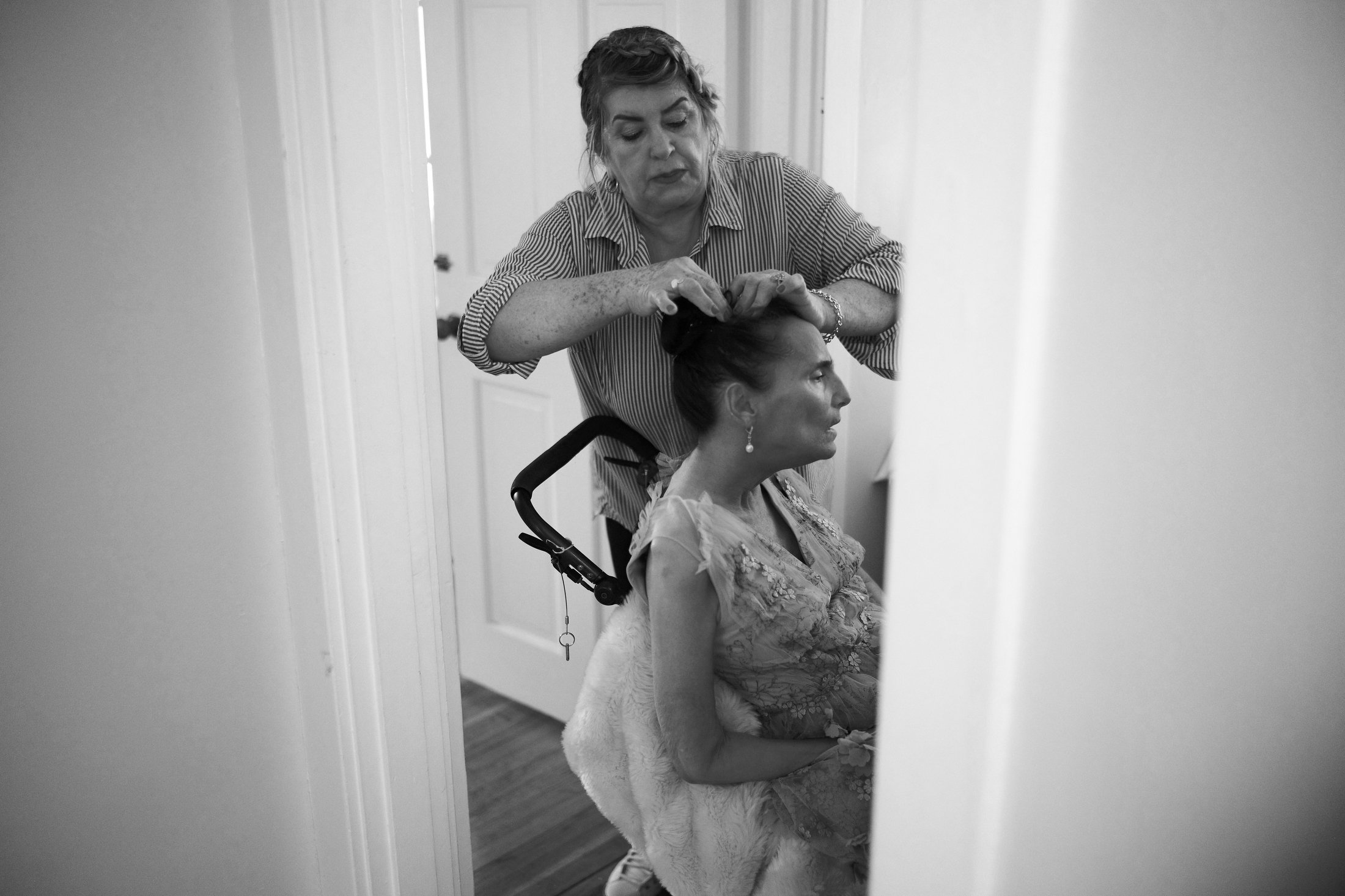A woman is sitting in a wheelchair with a woman styling her hair, both are indoors in a room with a door and wooden floor.