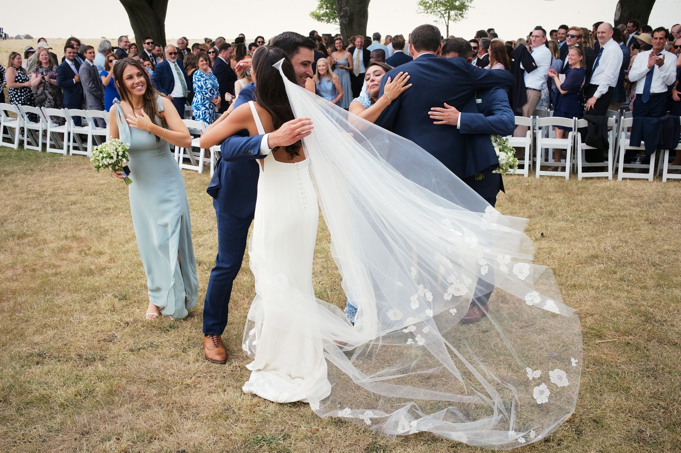 A bride in a white dress and veil hugging a groom in a navy suit, with a bridesmaid in a light blue dress holding a bouquet standing nearby, while many guests are clapping and smiling in the background at an outdoor wedding ceremony.