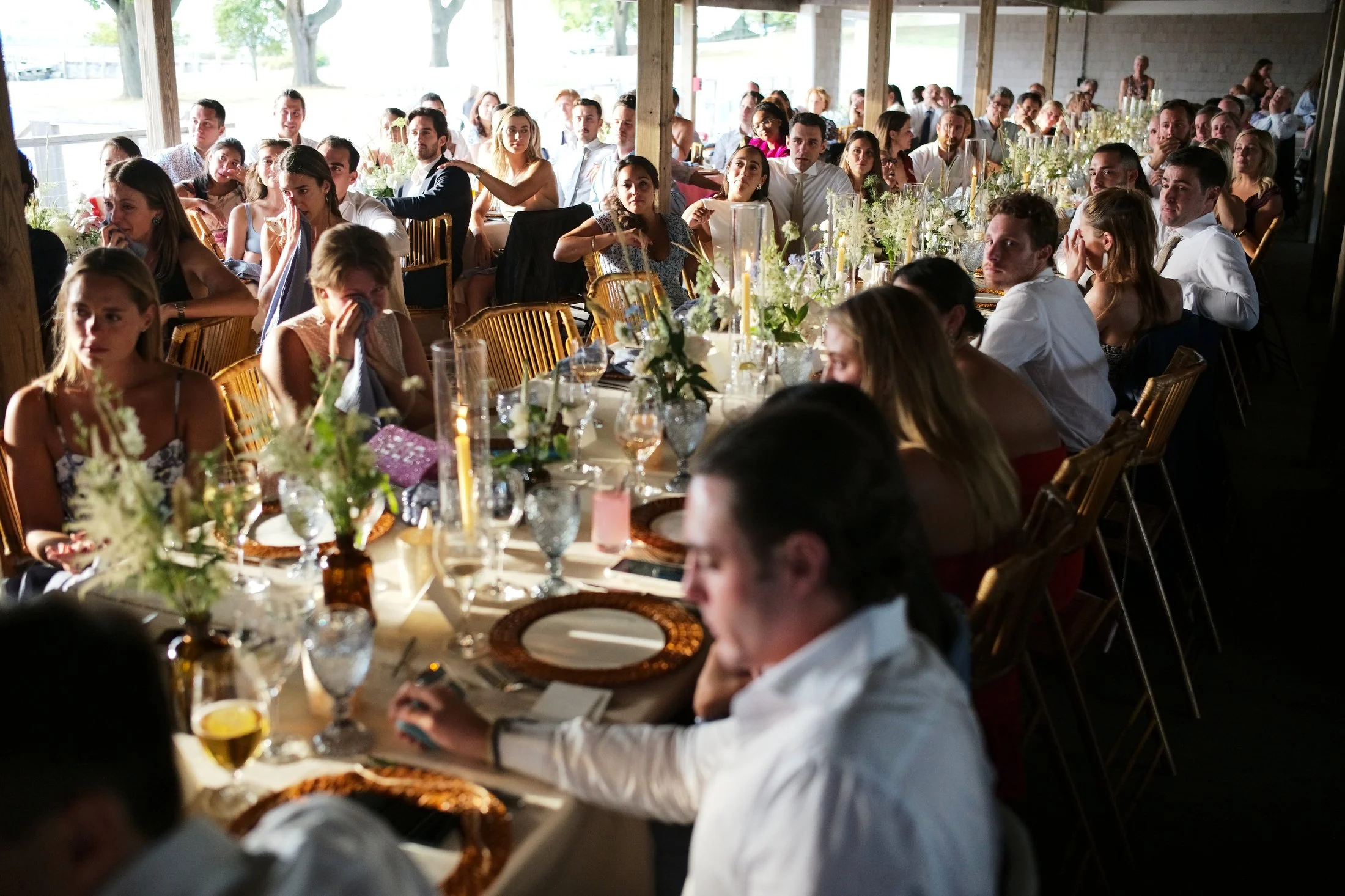 Guests seated at a long, decorated banquet table during a wedding reception or similar celebration, with floral centerpieces and candles, in a bright, open-sided venue.