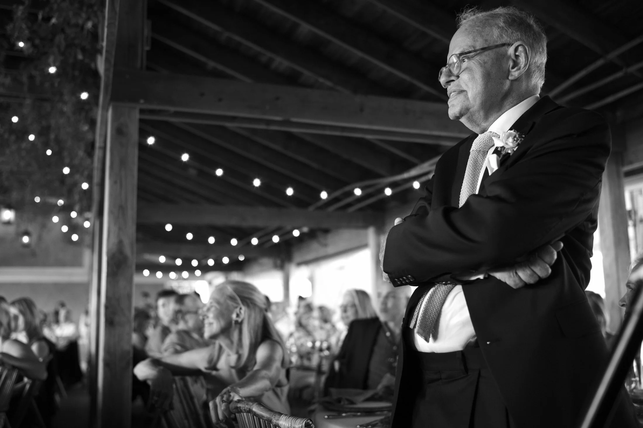 A man in a suit and tie standing with arms crossed, watching an event in a decorated venue filled with seated guests.