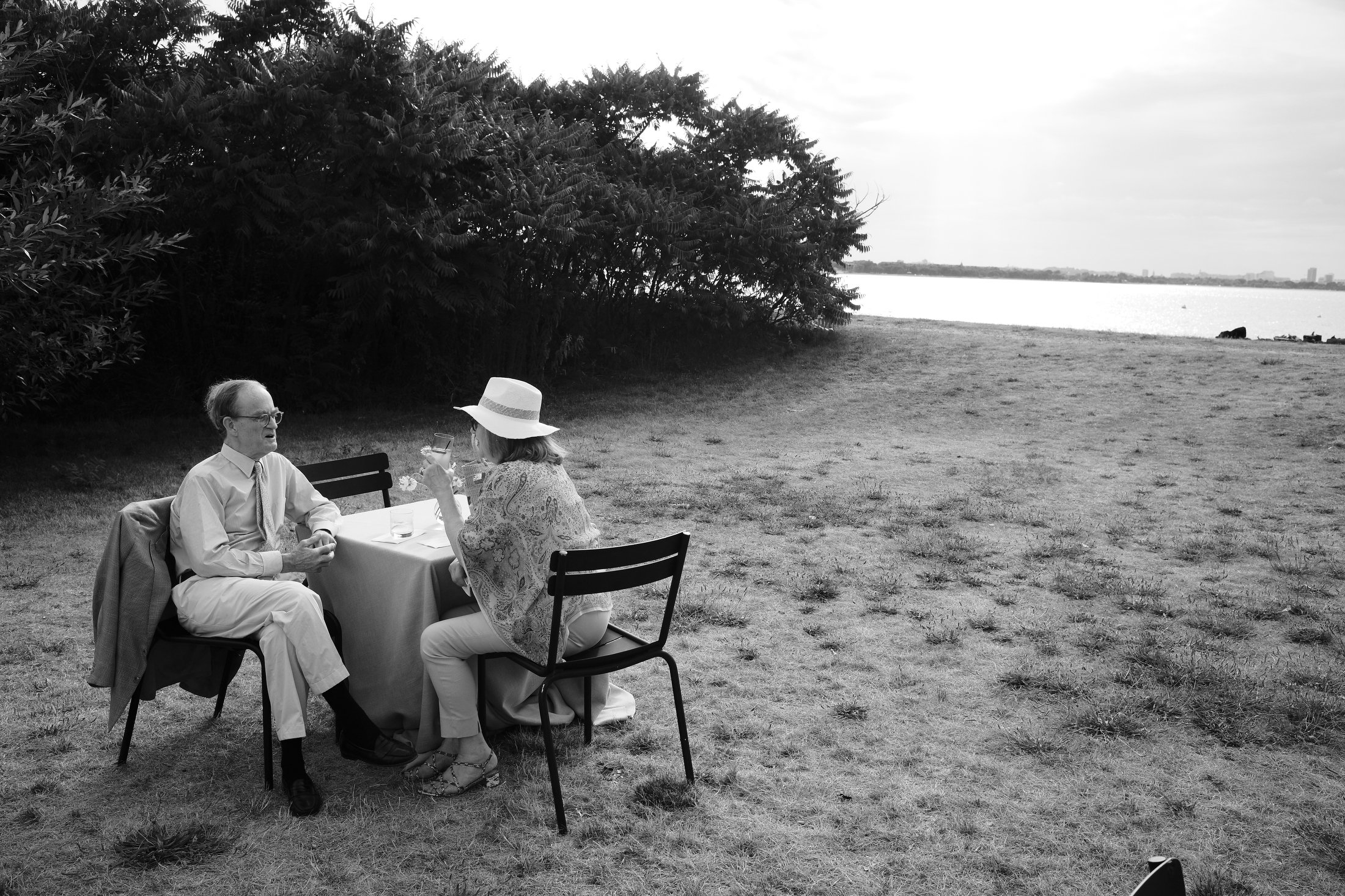 A man and woman sitting at a table outdoors near a body of water, having a conversation and holding glasses. The woman is wearing a wide-brimmed hat and patterned blouse, the man wears glasses and a tie. There is a jacket hanging on the back of the m