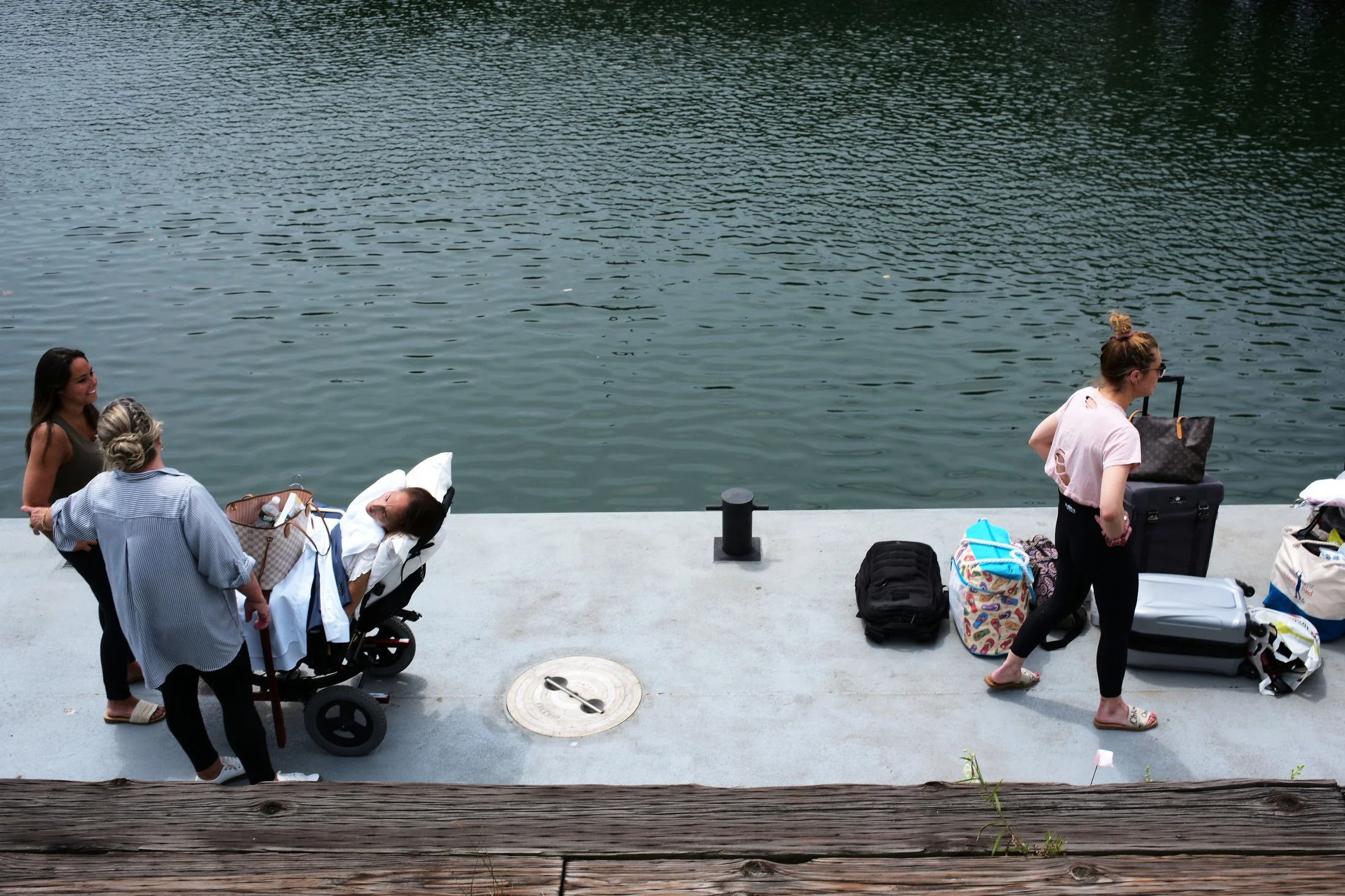 Four women with luggage and a stroller by a body of water, with two women talking and one woman facing away from the water