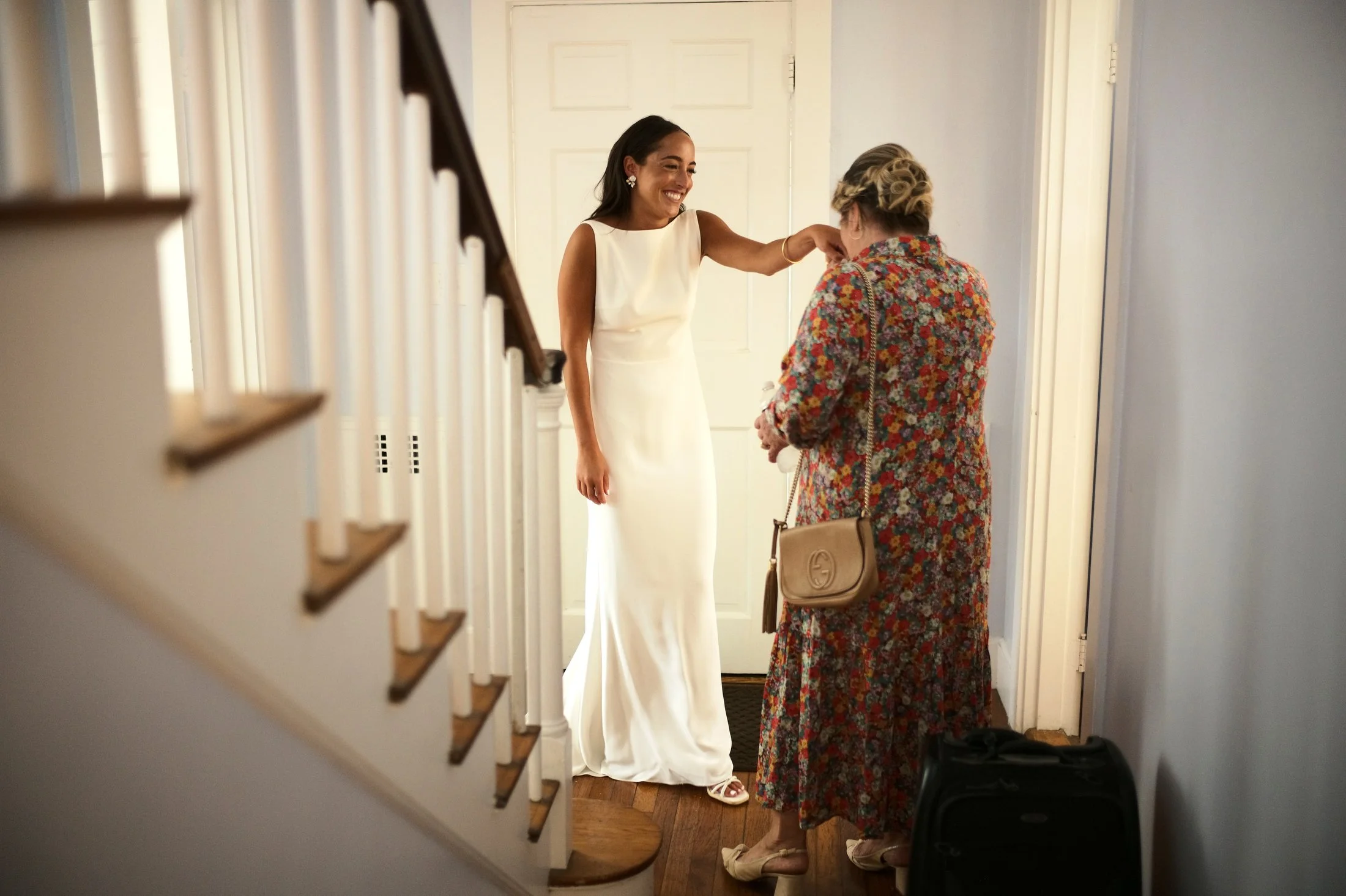 A woman in a white wedding dress smiling and sitting down as an older woman in a floral dress adjusts her necklace inside a house.