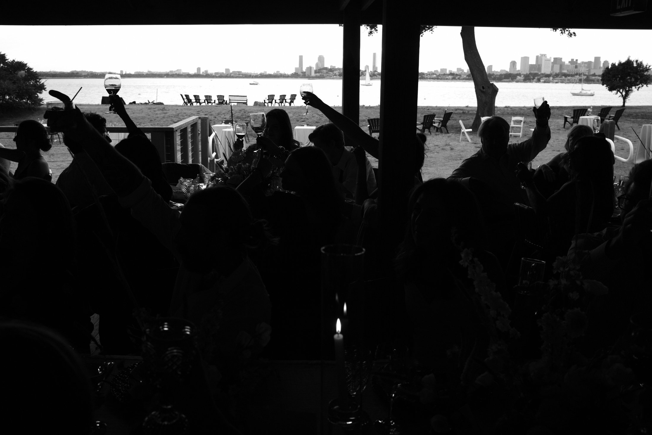 Silhouetted group of people raising glasses at a dinner party under a pavilion, with an outdoor setting of chairs, tables, and a waterfront with sailboats and a city skyline in the background.