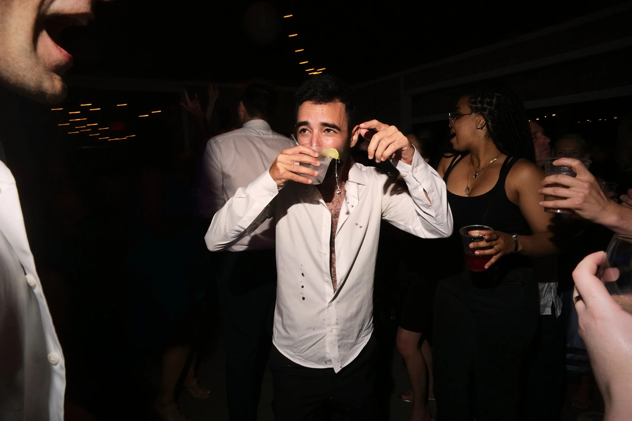 Group of people at a party with drinks, including a man in the center with a white shirt, holding a drink with a lime wedge, smiling and dancing.