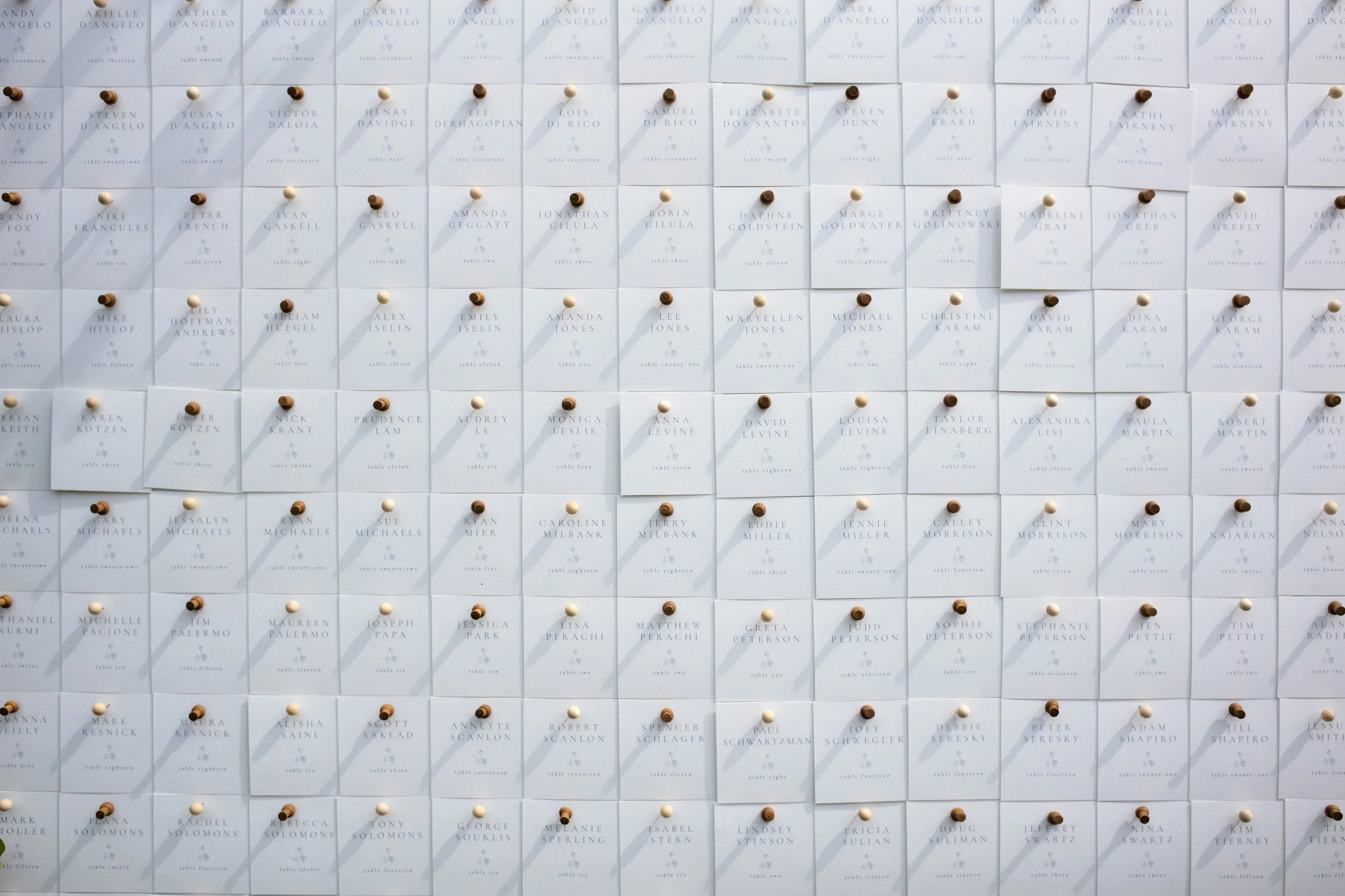A wall display with numerous white sheets of paper pinned with brown and white thumbtacks. Each sheet has a name and table assignment printed on it, arranged in a grid pattern.