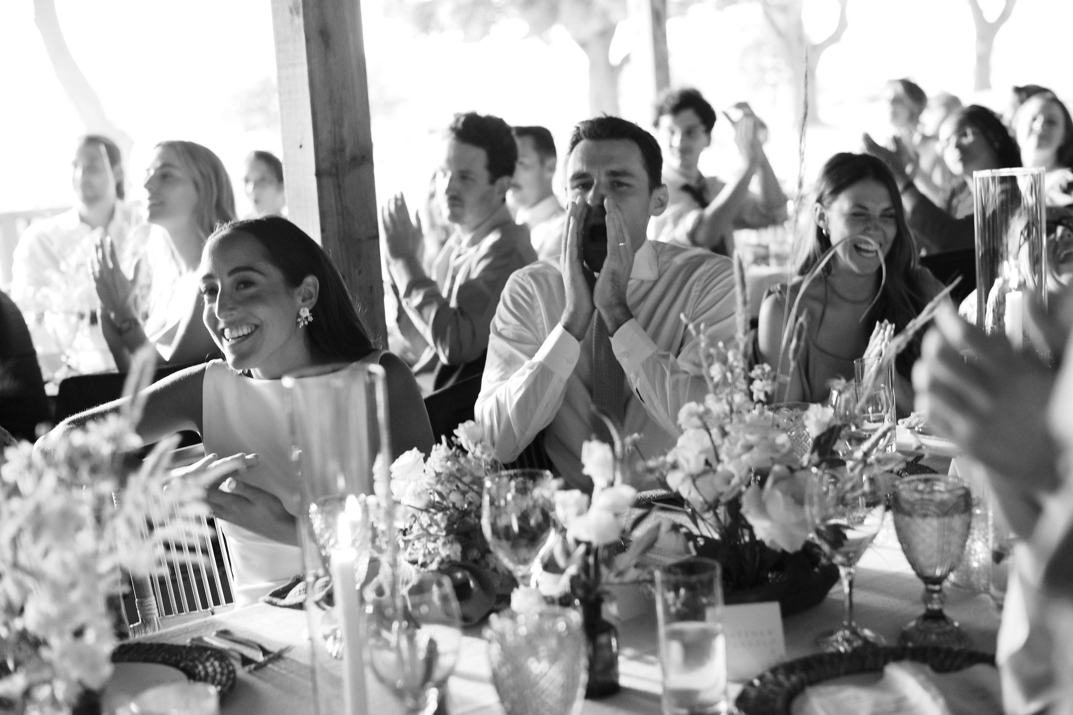 A group of people sitting at a table during a celebration, smiling and clapping, with some covering their mouths or reacting excitedly, decorated with flowers and glassware.