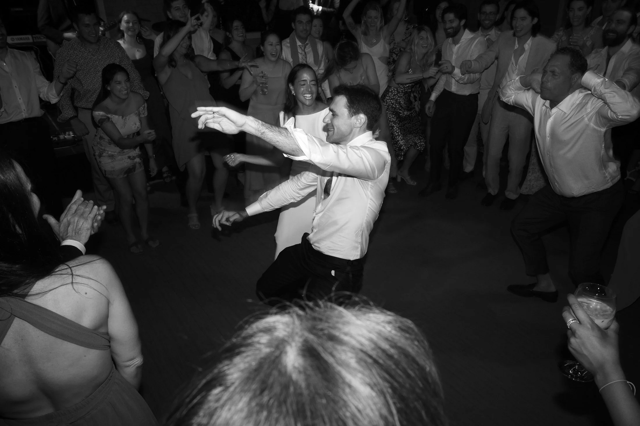 People dancing and celebrating at a party or wedding reception, with a man and woman in the center dancing closely surrounded by smiling guests.