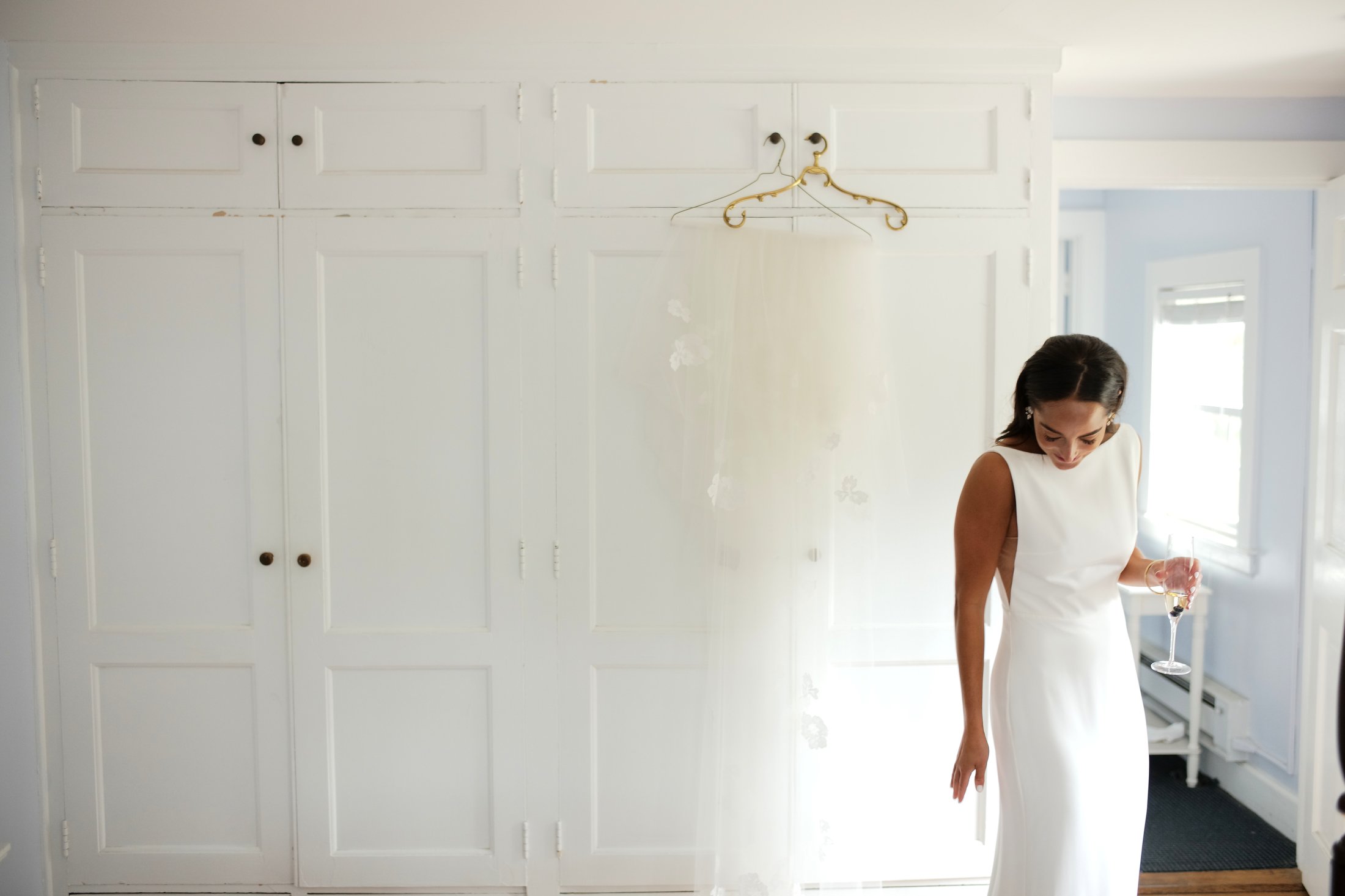 Woman in white dress holding a glass of wine, looking down, standing next to a large white closet in a bright room.