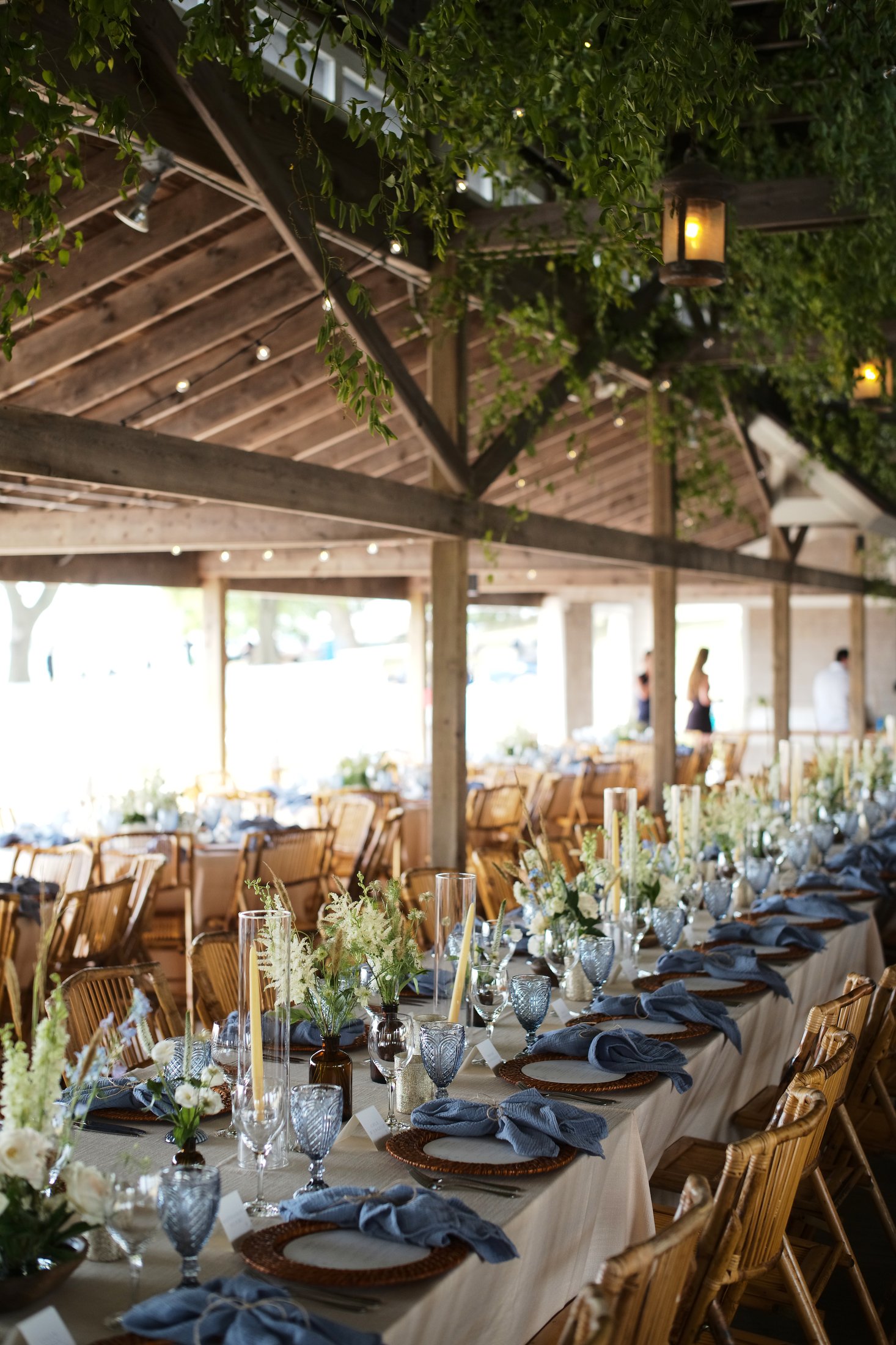 Long banquet table with floral centerpieces, blue napkins, and place settings in an outdoor rustic covered venue with wooden beams, hanging lanterns, greenery, and some people in the background.