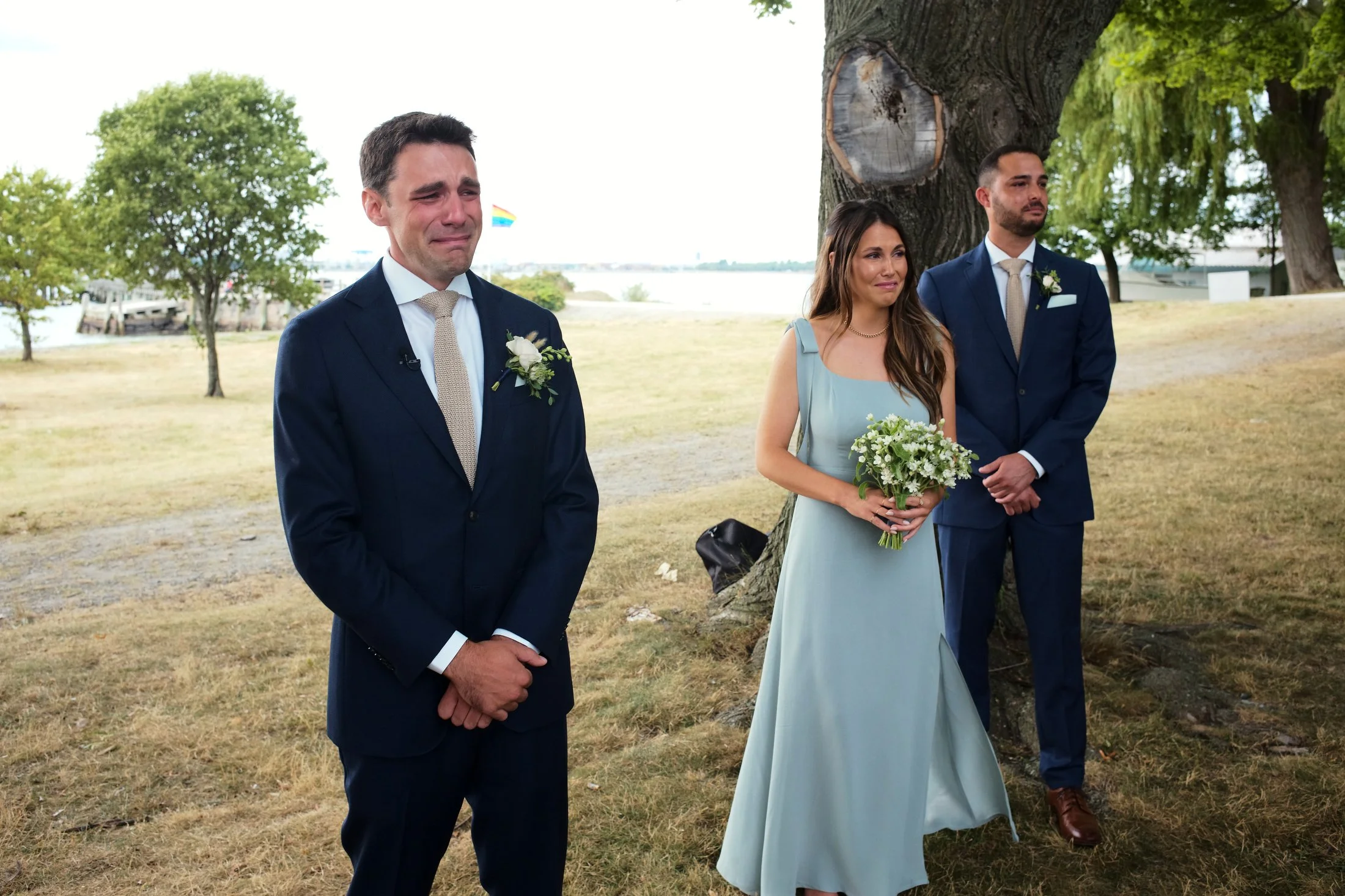 A wedding ceremony outdoors under a large tree. The bride is holding a bouquet of white flowers, standing next to a man in a dark suit who is emotional, with tears and a heartfelt expression. Another man in a dark suit stands nearby. The scene is set