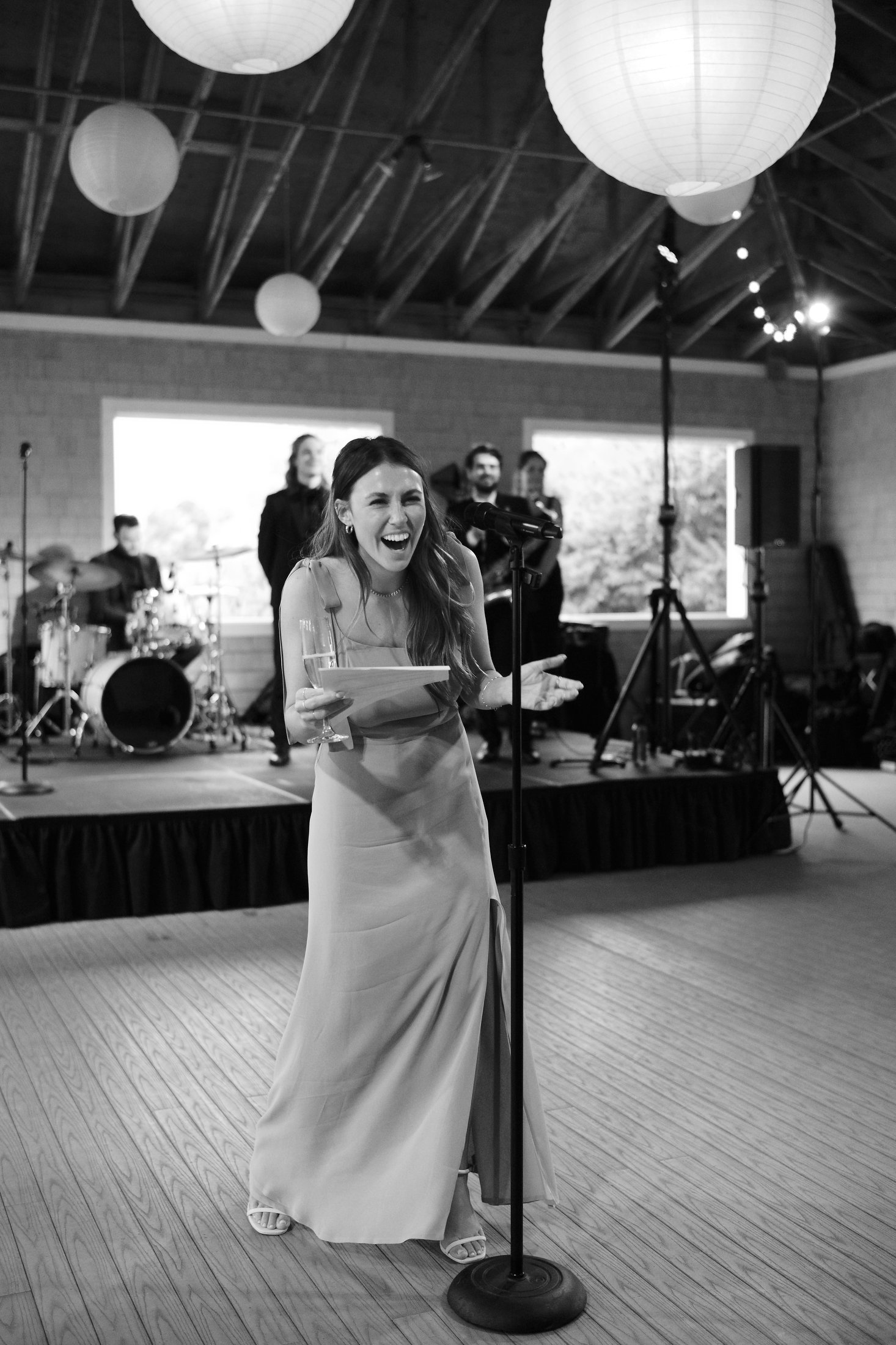 A woman in a formal dress is laughing while speaking or singing into a microphone, with a band playing in the background at an indoor event or celebration.