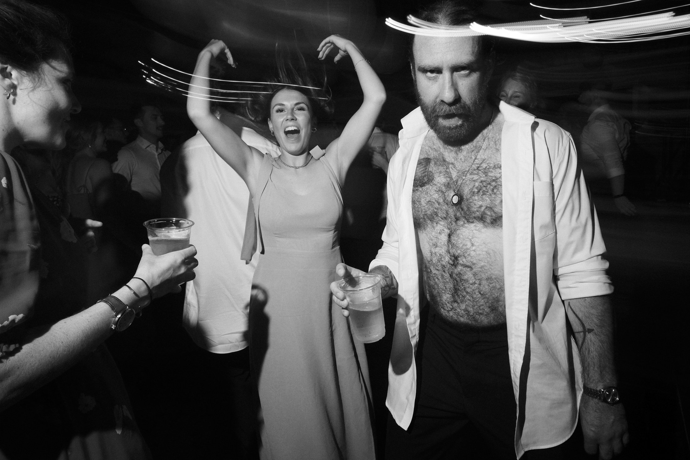 People enjoying a lively party, with a woman dancing and smiling, a man with a beard and chest hair holding a drink, and others in the background with drinks, in black and white.