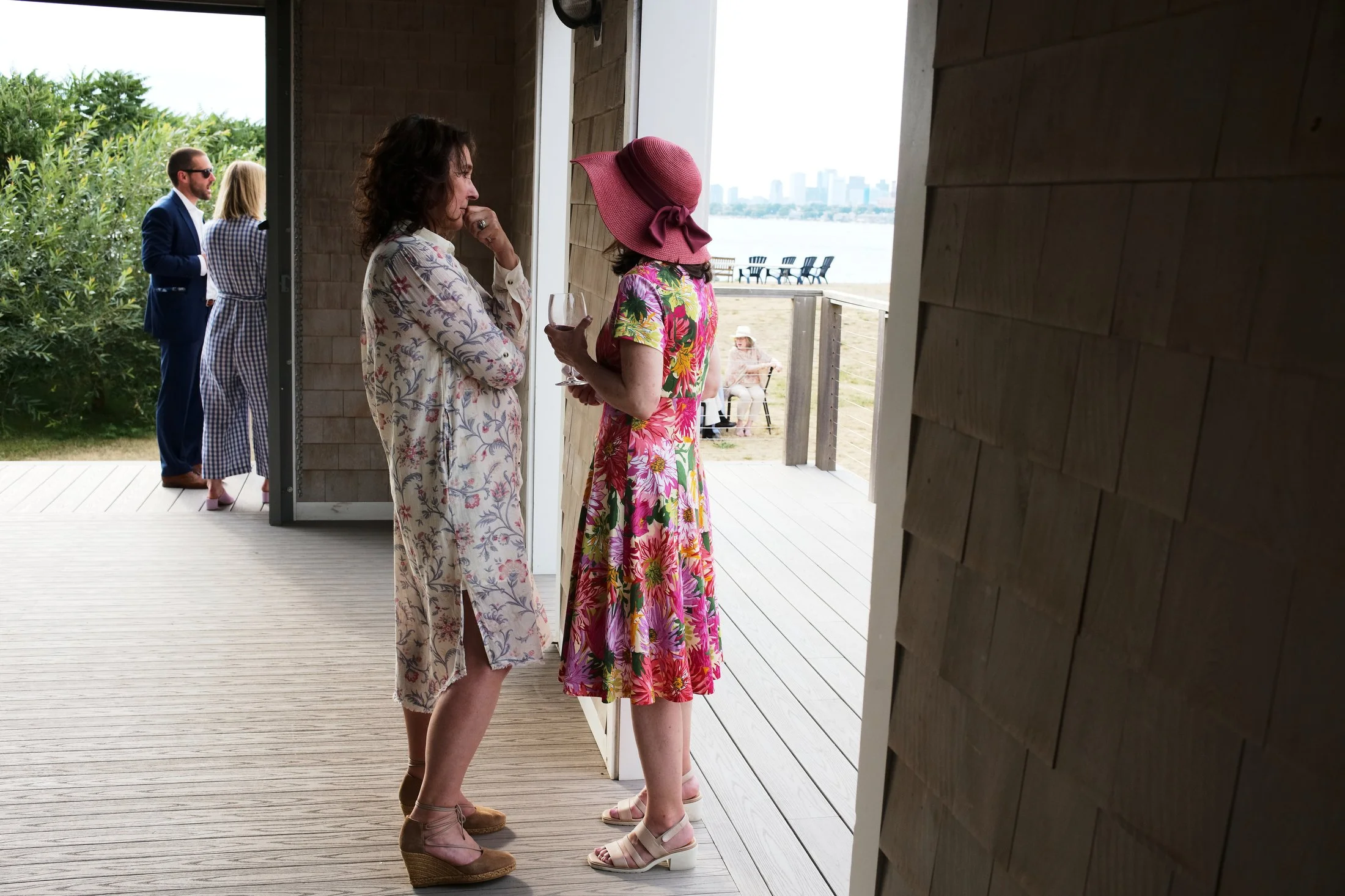 Two women having a conversation inside a house with a deck, with three people outside near the water in the background.