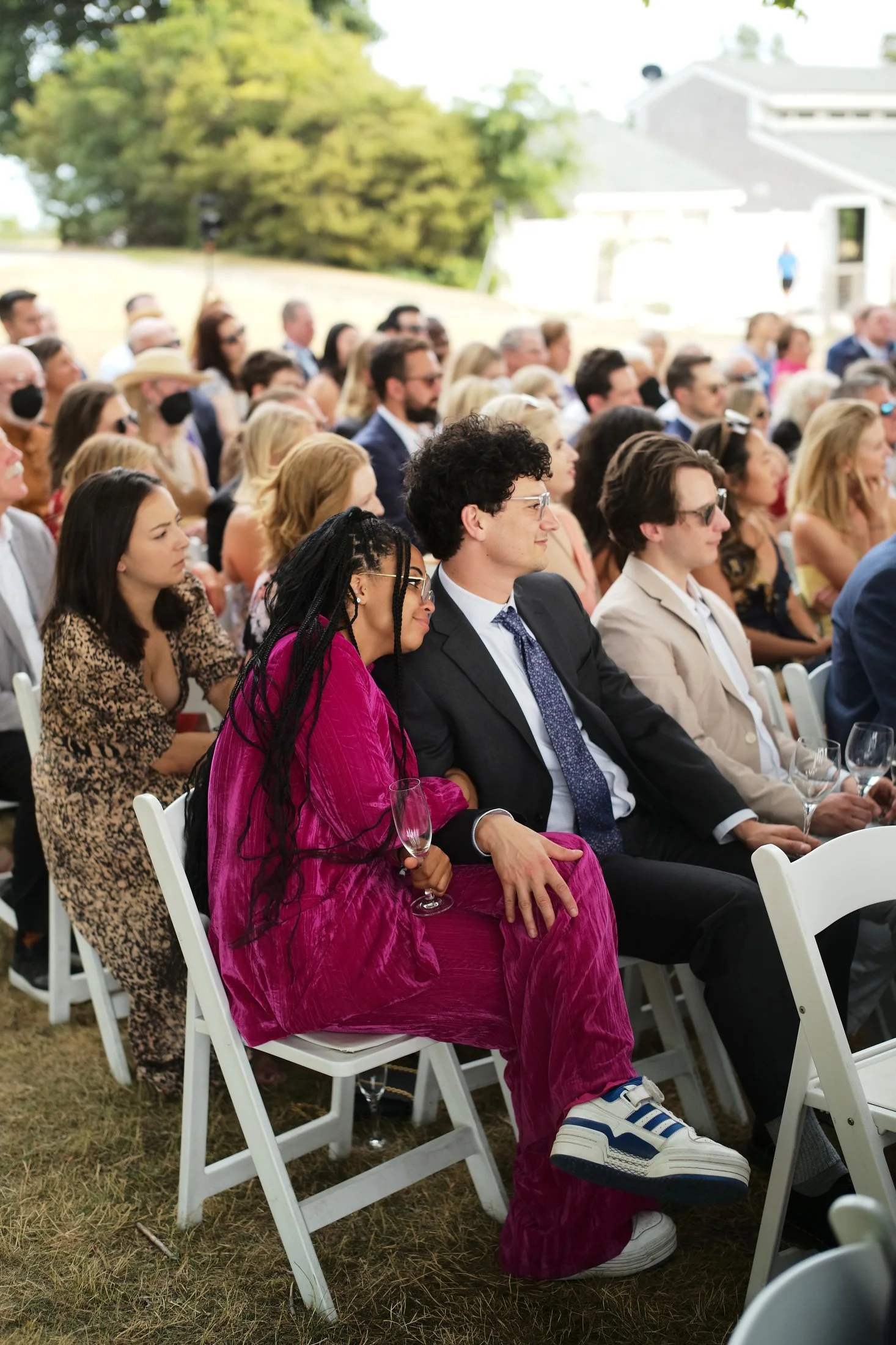 People attending an outdoor event, sitting in white chairs, some holding wine glasses, dressed in formal and casual attire, with trees and a building in the background.