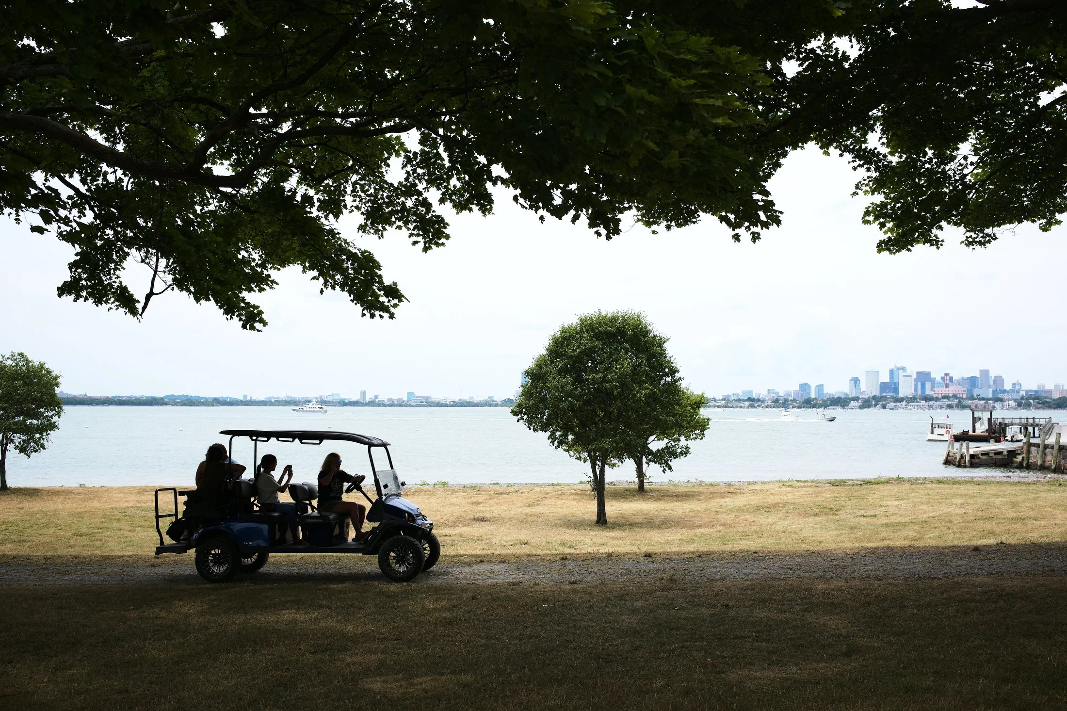 A group of people riding in a golf cart along a lakeside park with trees, water, and city skyline in the background.