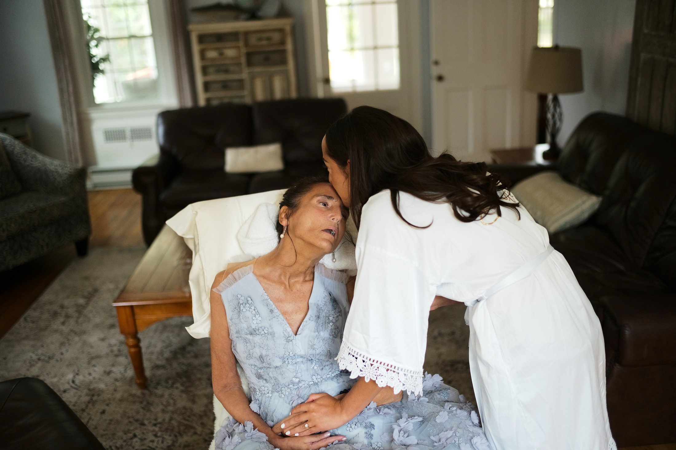 A woman lying in bed and an older woman with white hair and dressed in a light blue gown are sharing an intimate moment in a living room.