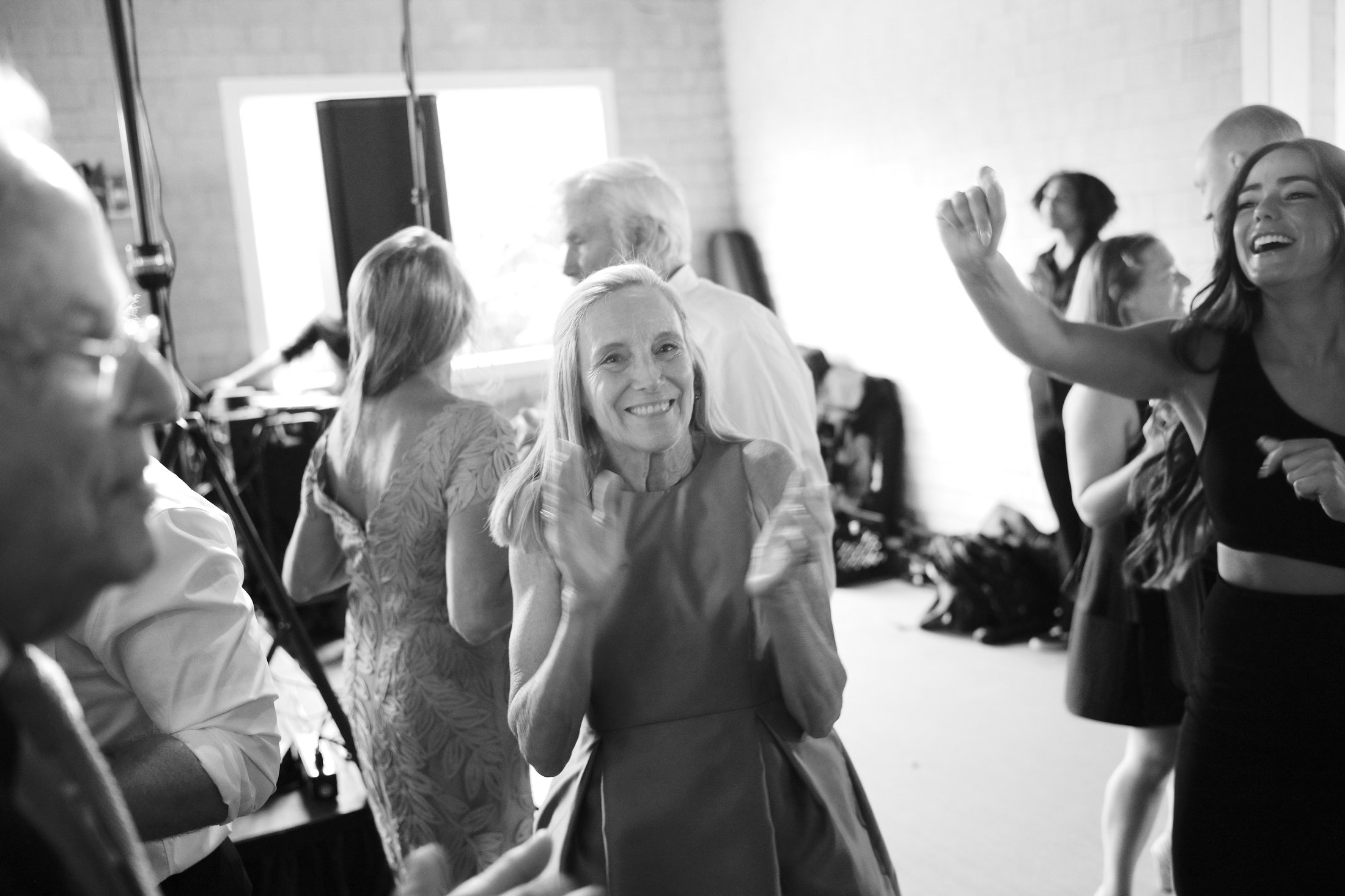A woman with long hair in a dress is smiling and dancing with others at a gathering or party.