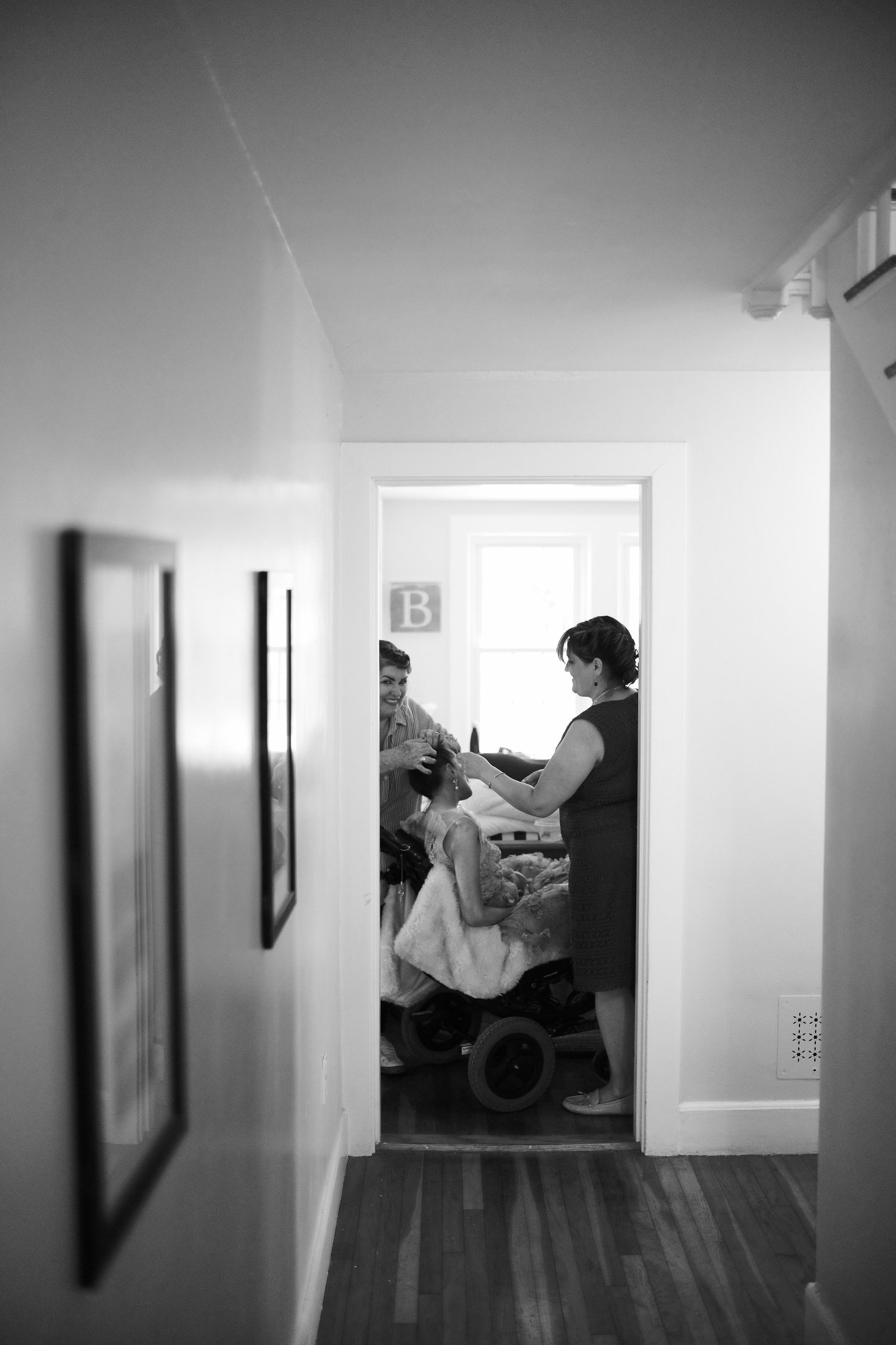 Two women and a young girl in a room, with one woman doing the girl's hair while the other woman holds a toy, seen through a doorway in a black-and-white photo.