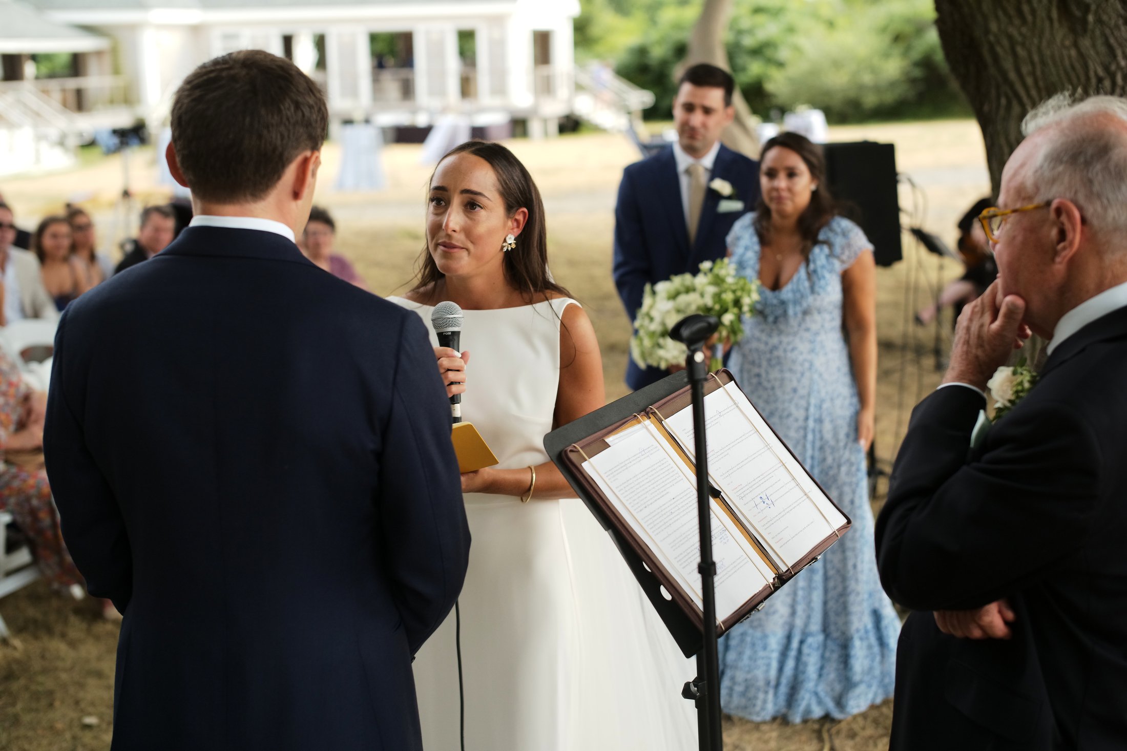 A woman in a white wedding dress appears to be reading her vows into a microphone during an outdoor wedding ceremony, with the groom in a dark suit facing her. In the background, a woman in a blue dress and a man in a suit and tie stand near a tree, 