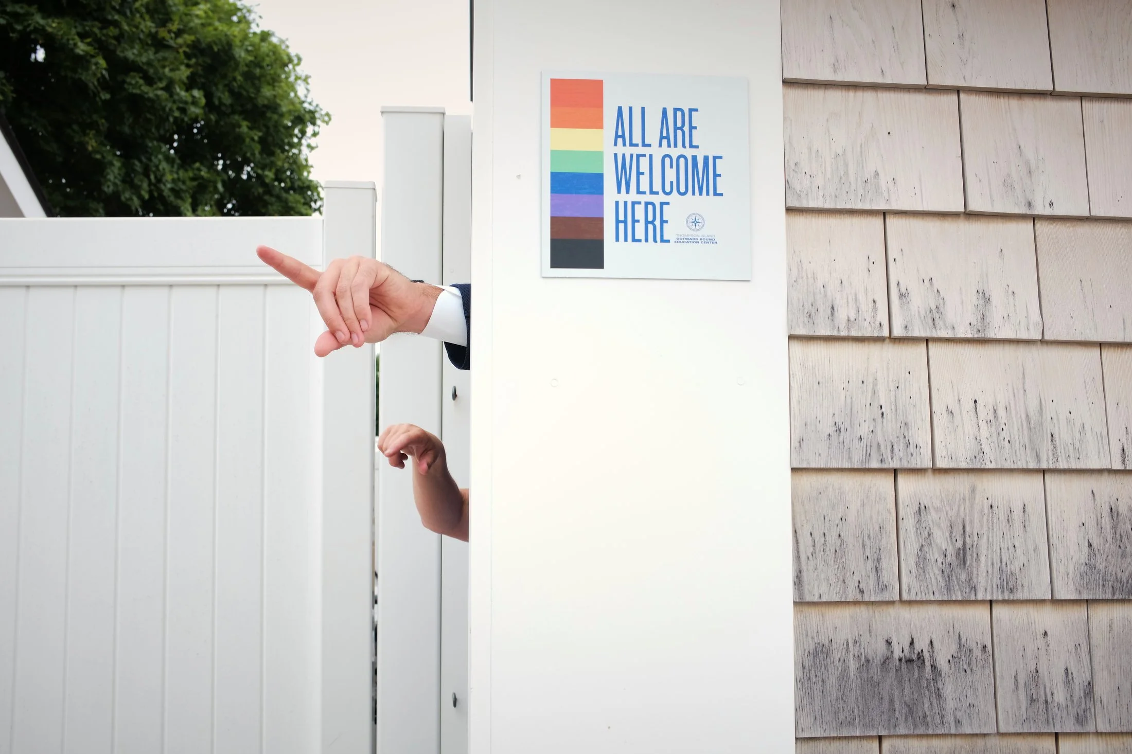 Two hands, one with a suit sleeve, are giving a thumbs-up and a fist bump through a white fence. There is a sign on the fence that says "All Are Welcome Here" with a rainbow-colored stripe.