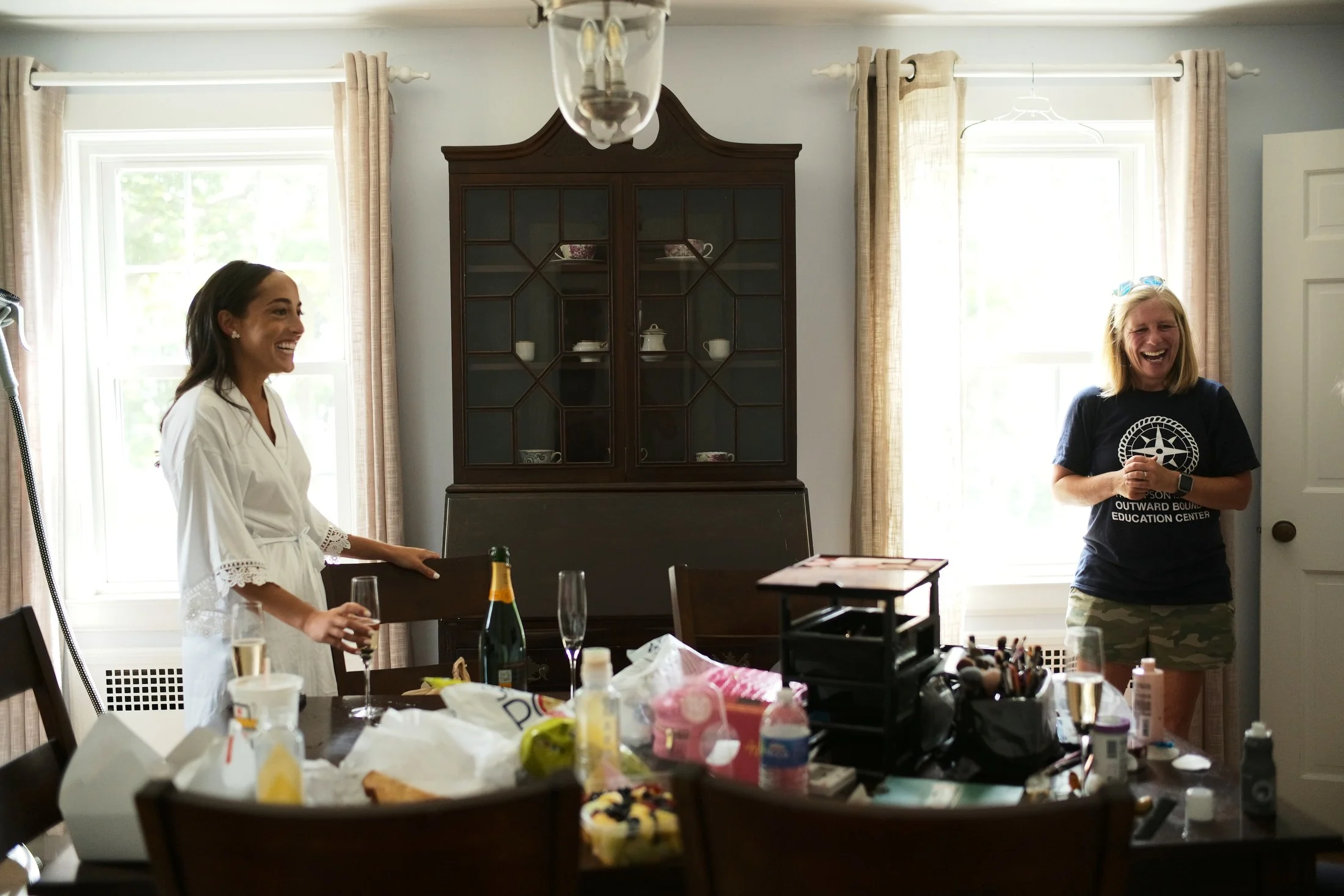 Two women are in a dining room, smiling and laughing. One woman is standing near a table, holding a glass of champagne, while the other is standing near a window, wearing a black t-shirt and shorts. The table has various items on it, including bottle