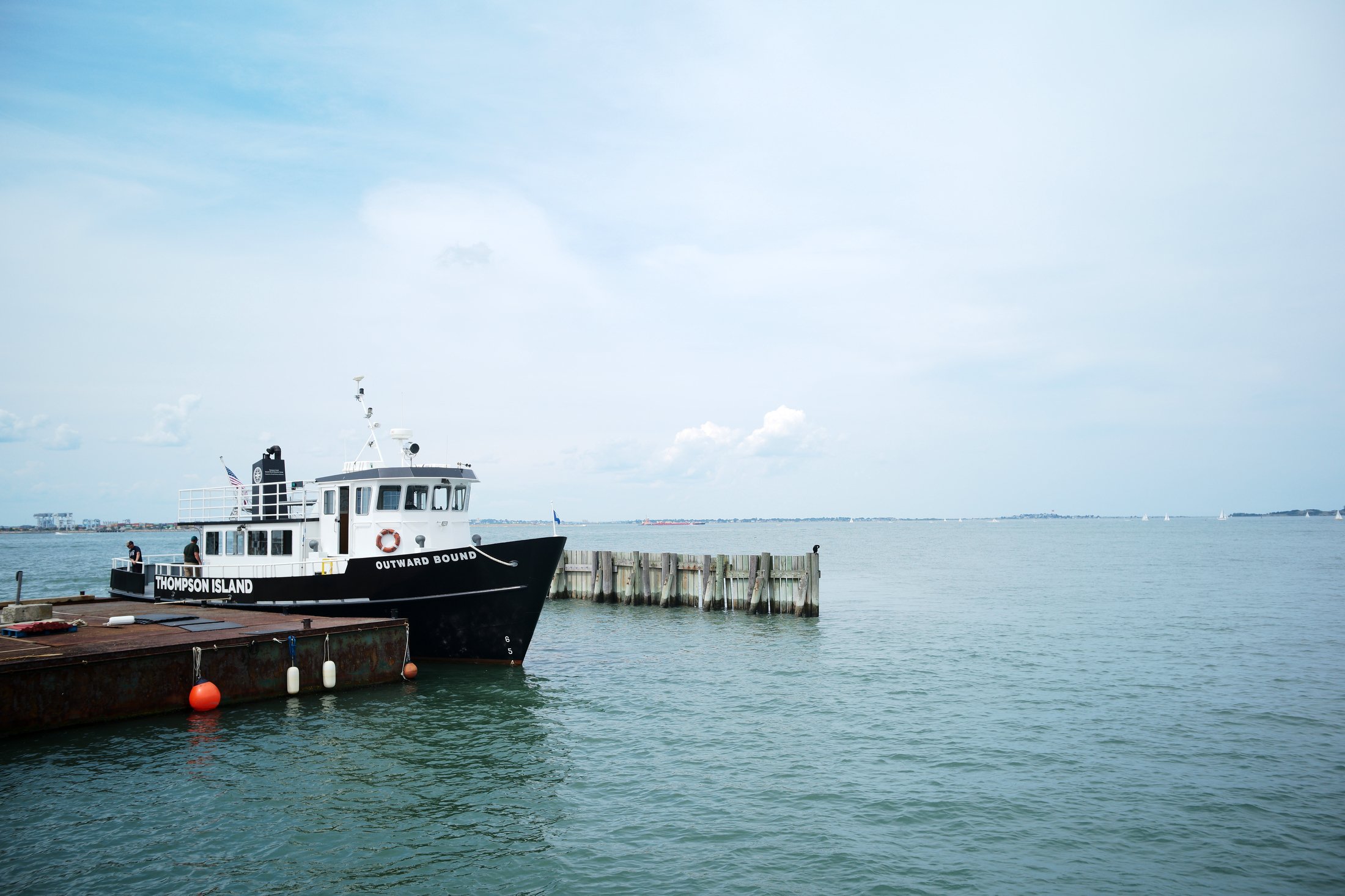 A black and white boat named 'Thompson Island' docked at a pier on calm water with a cloudy sky overhead.