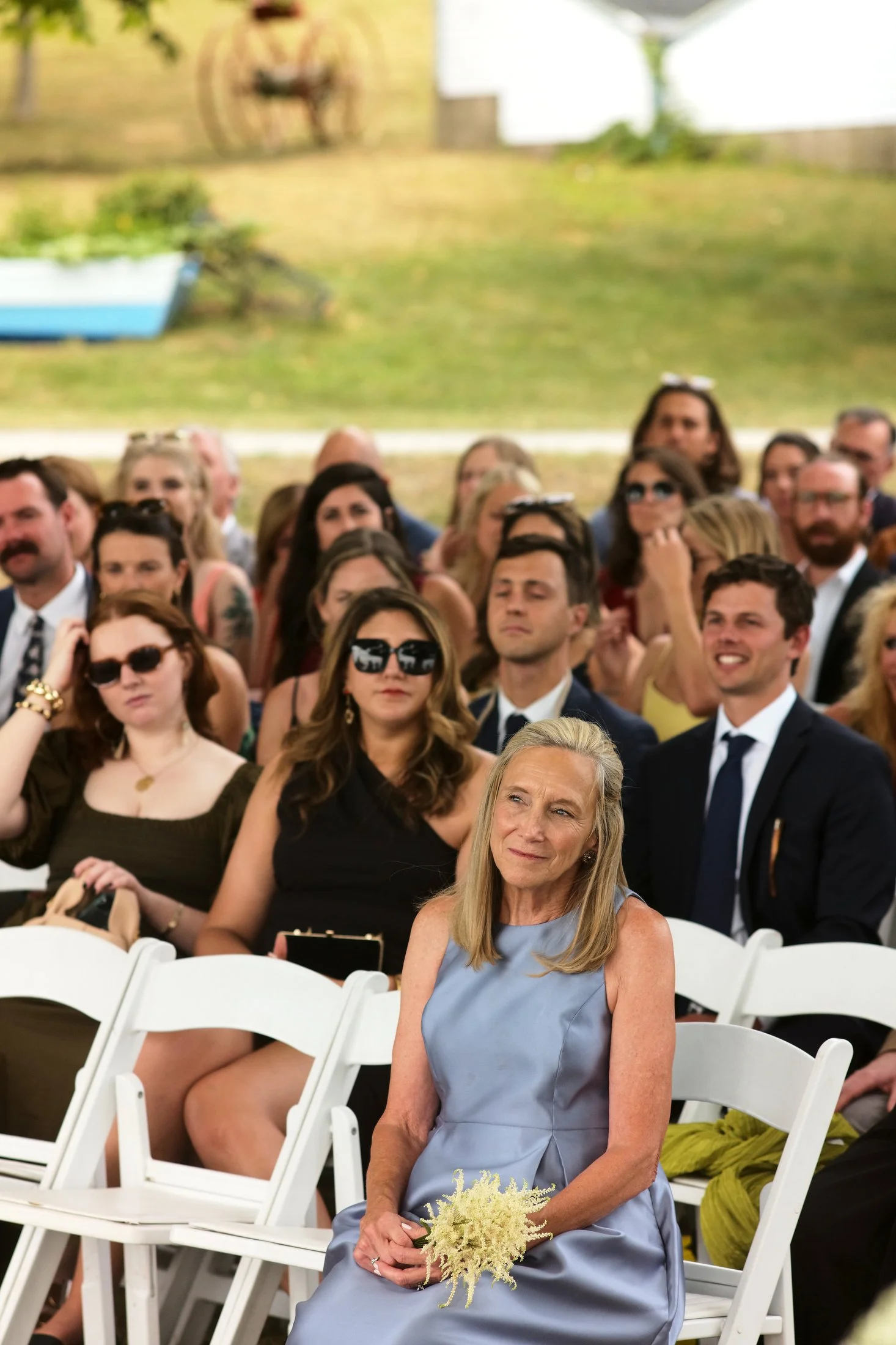 An elderly woman in a light blue dress sitting on a white folding chair at an outdoor wedding ceremony, holding a small bouquet of flowers, with a large group of seated guests behind her.