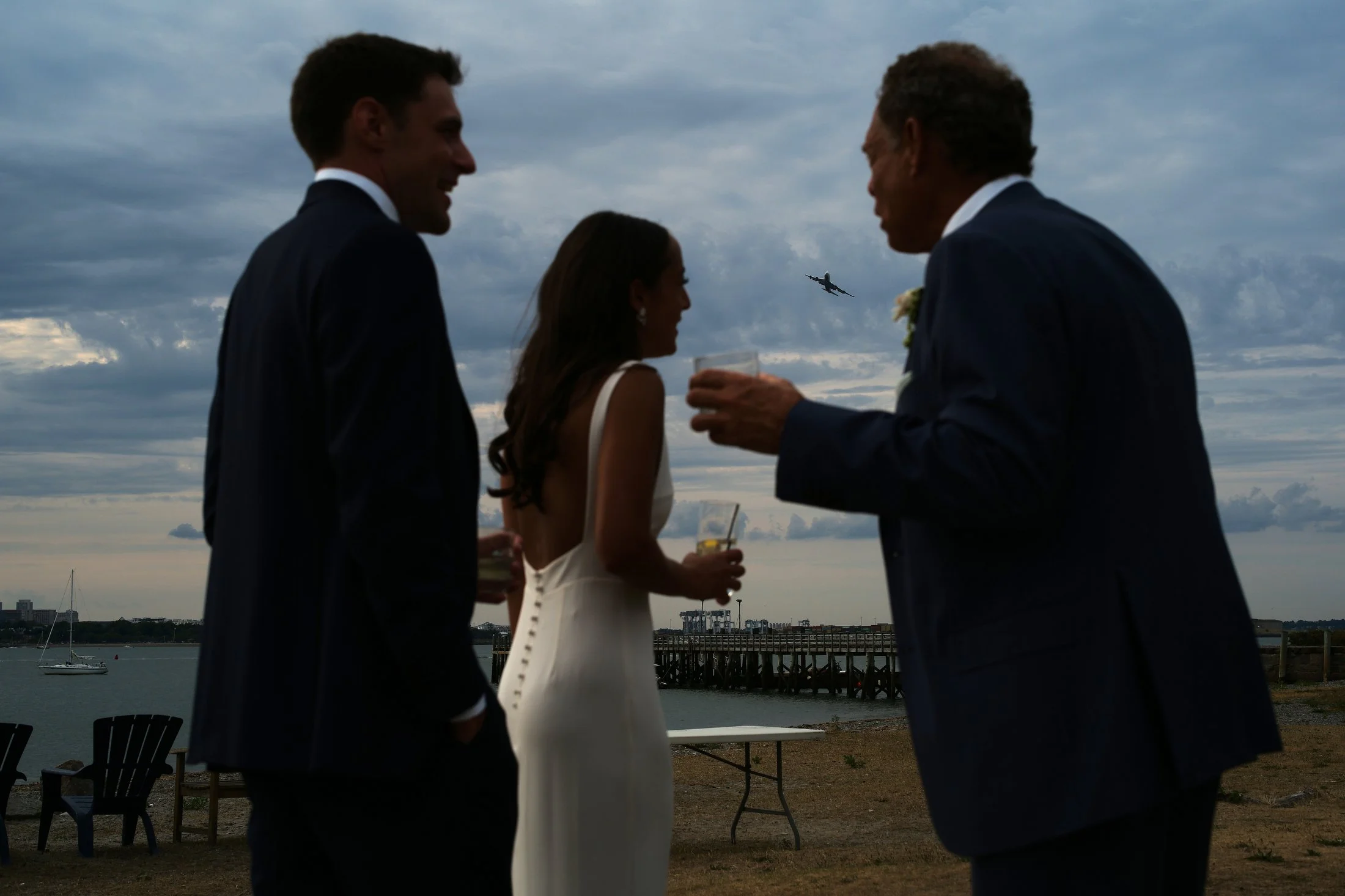 Four people in formal attire are socializing outdoors near the water during dusk, with an airplane flying in the cloudy sky in the background.