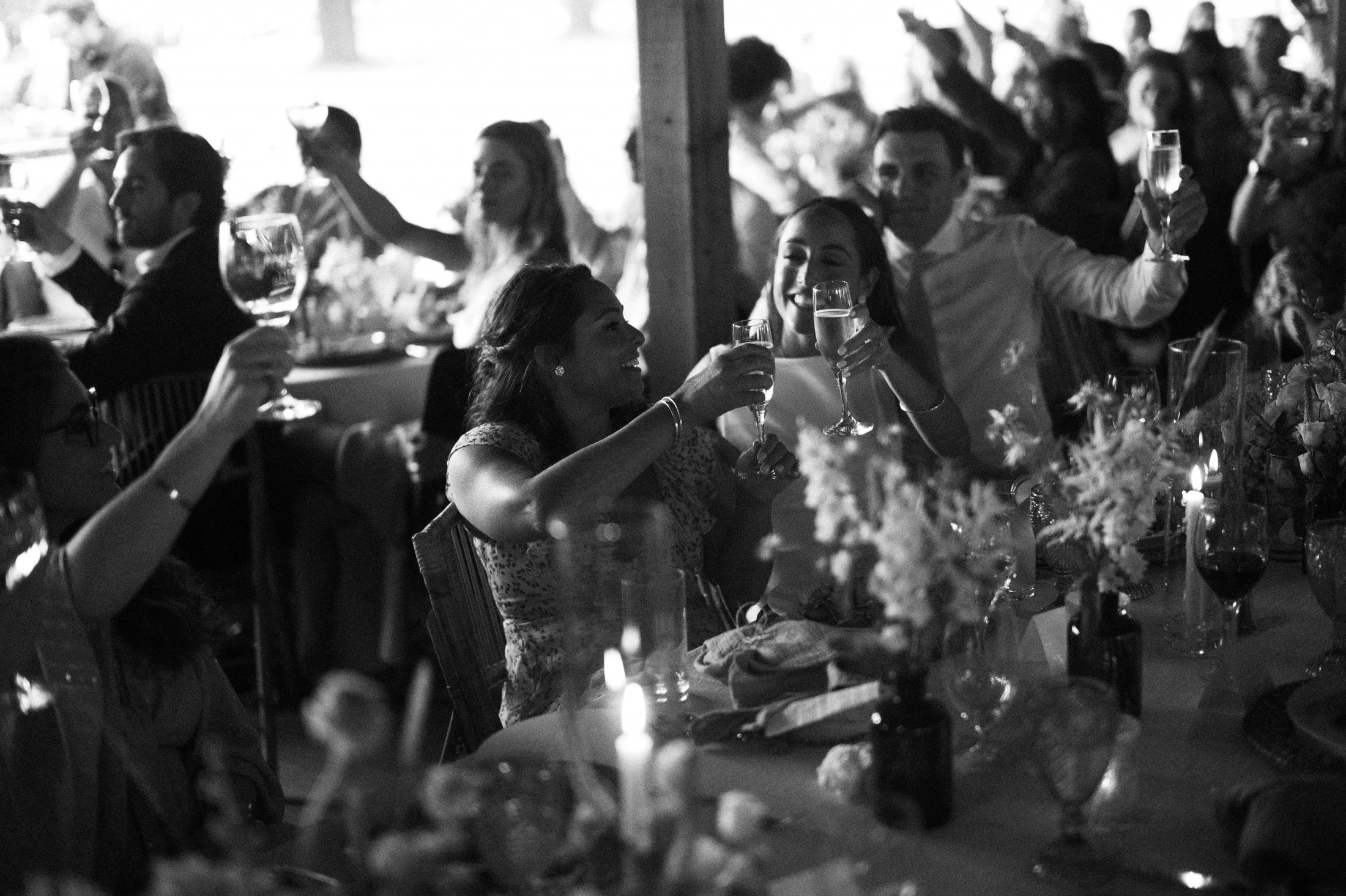 People raising glasses in a toast at a lively banquet or celebration, in a formal setting with decorated tables and floral centerpieces, black and white photograph.
