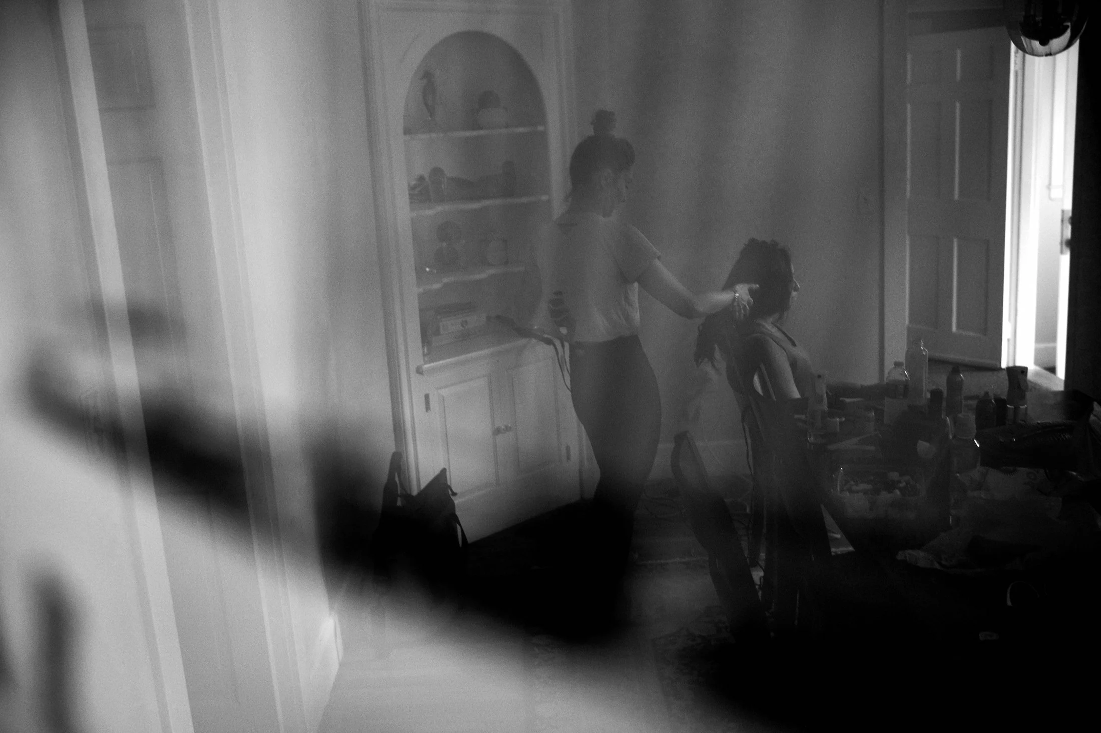 A woman is sitting in front of a mirror, getting her hair styled by another woman in a room filled with makeup and beauty products, with a door doubled open in the background.