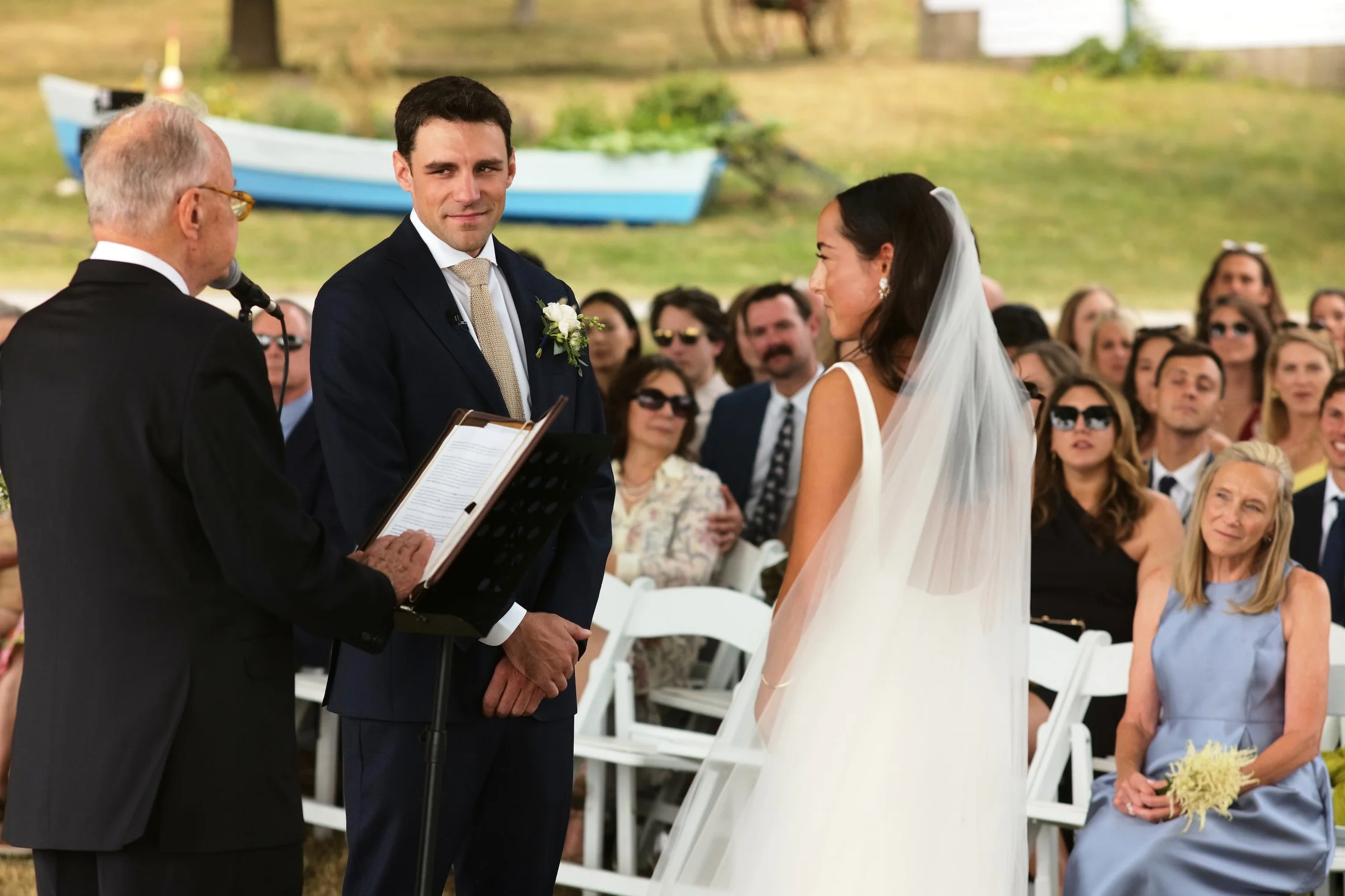 Groom and bride standing at an outdoor wedding ceremony, with officiant reading from a book, guests seated in the background.