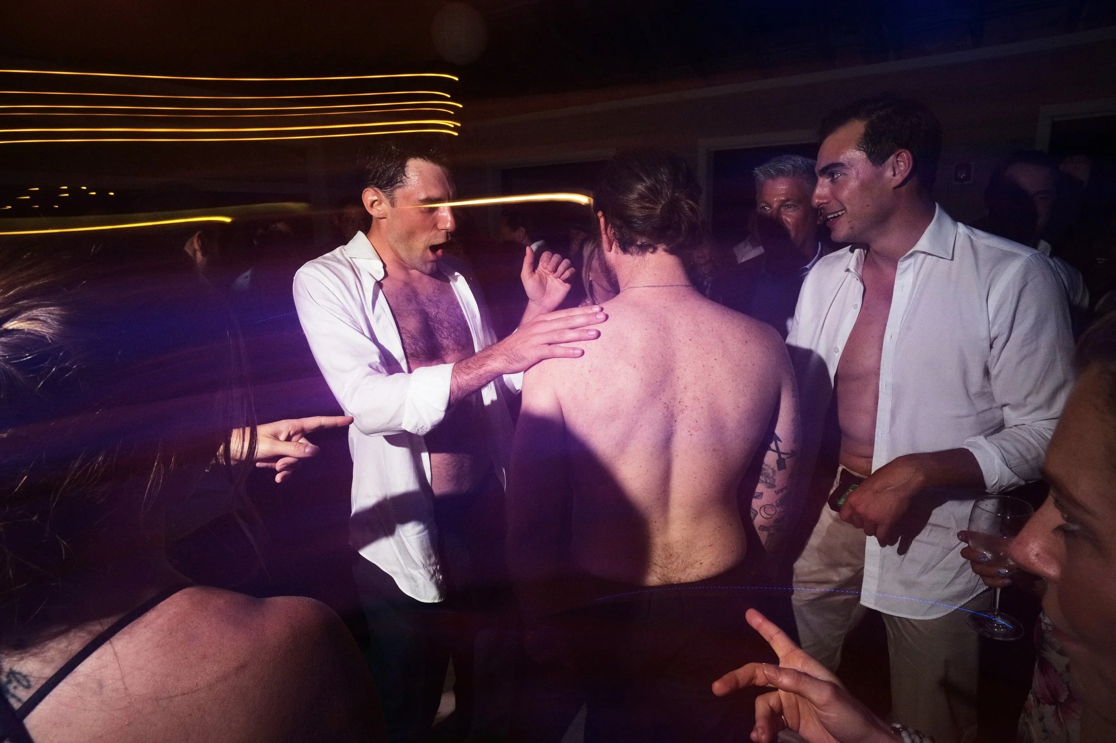 Group of men and women at a party, with one man shirtless, others wearing open shirts, in a dimly lit room with streaks of light from movement.