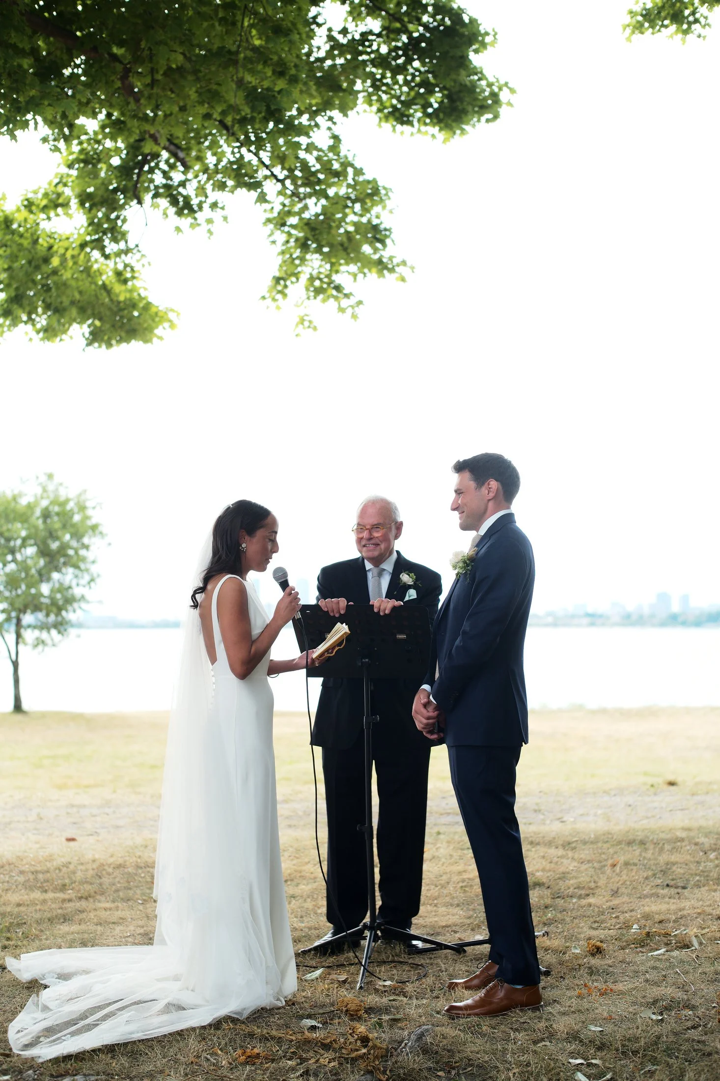 A bride and groom exchanging vows during an outdoor wedding ceremony by a lake under a tree, with an officiant standing between them