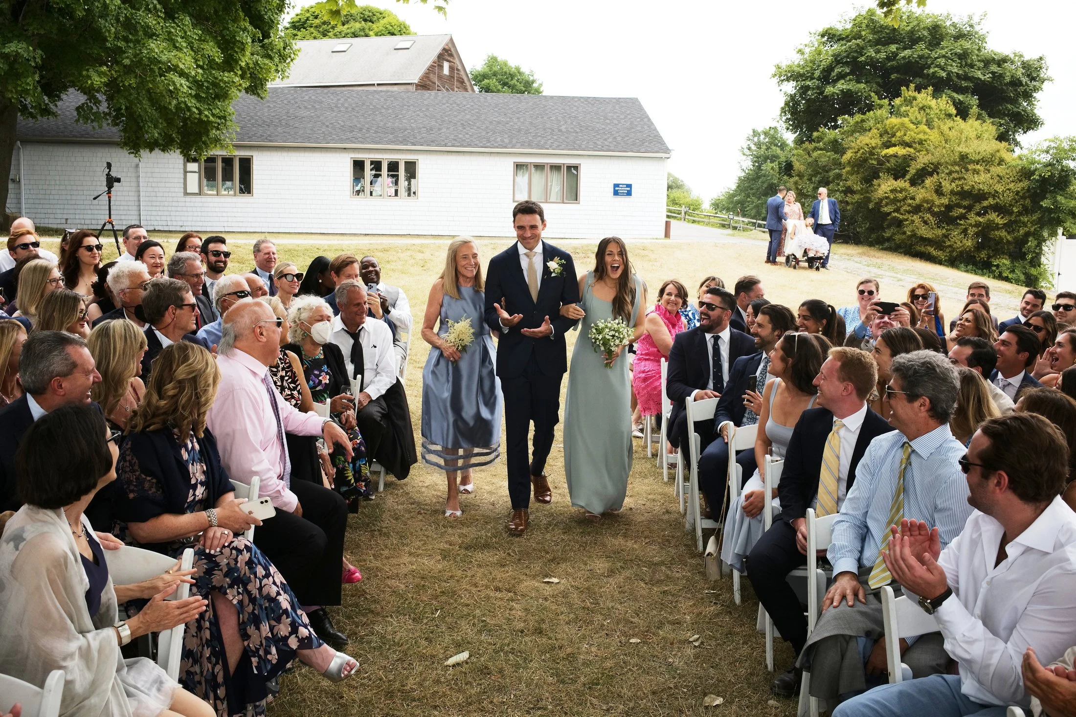 Bride and groom walking down the aisle at an outdoor wedding ceremony. Guests seated on both sides, watching and smiling.