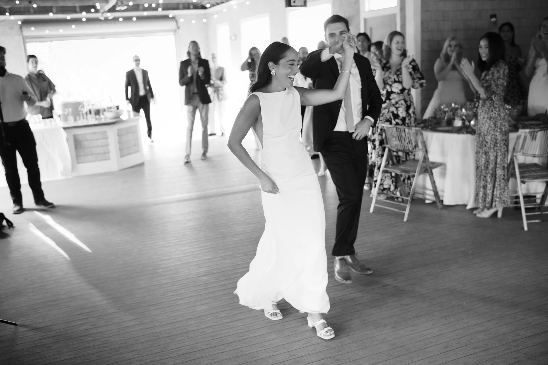 A woman in a white dress and a man in a suit dancing at a wedding reception with guests clapping and smiling in the background.
