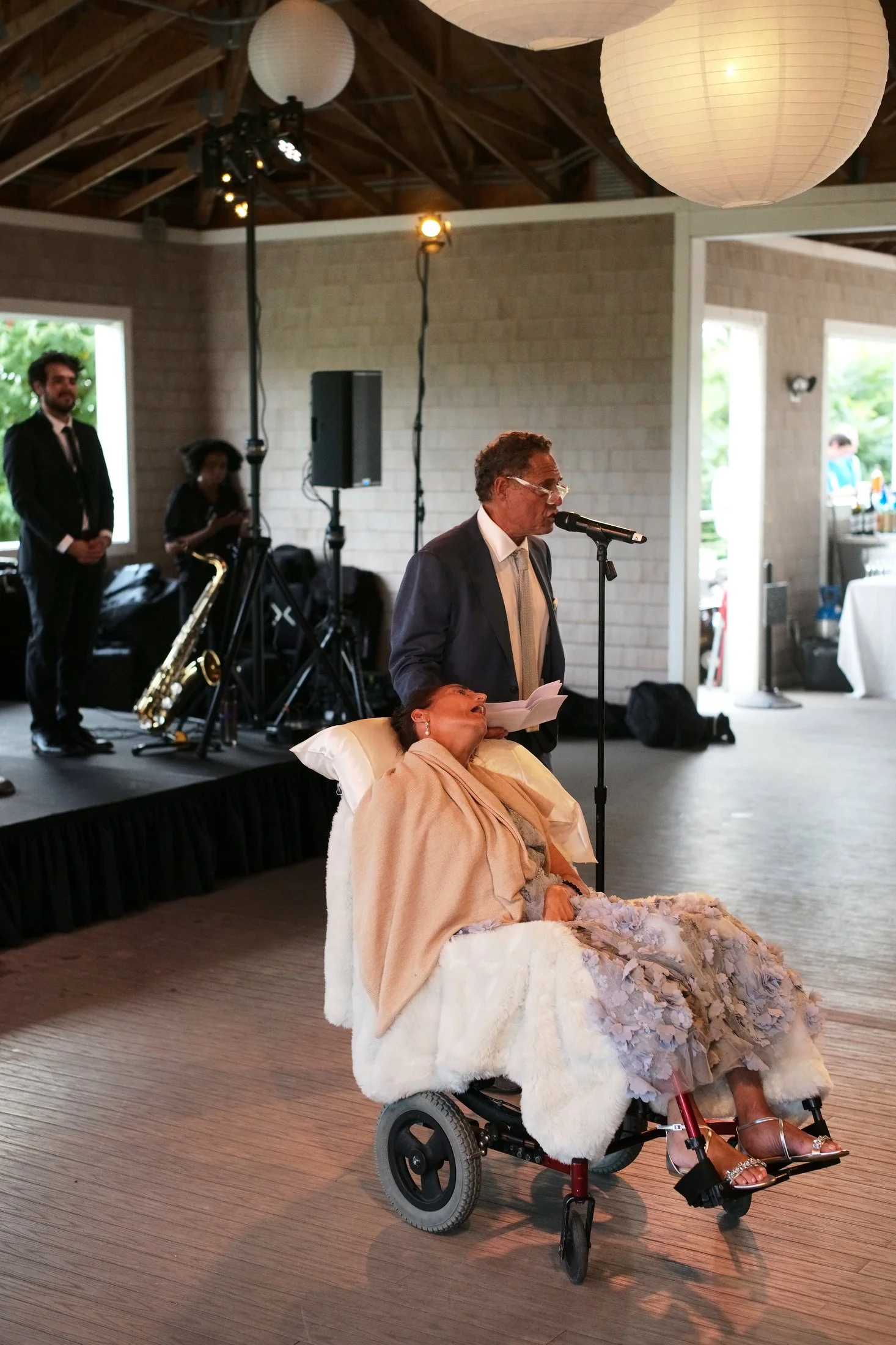 An elderly woman in a wheelchair, dressed in an elegant gown and shawl, is being accompanied by a man in a suit speaking into a microphone at a formal event. The woman appears to be asleep or resting her head back, with head tilted backward and eyes 