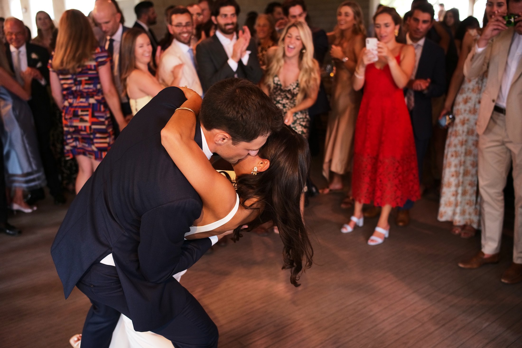 A newlywed couple shares a kiss during their wedding dance as guests gather around watching and taking photos.