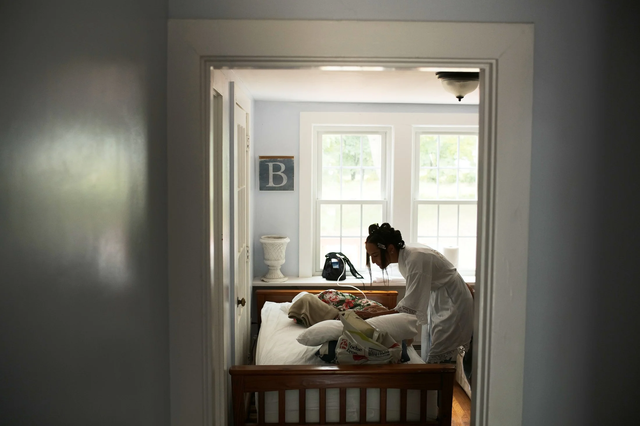 A woman in a white dress is tidying up a bedroom with a wooden bed frame, pillows, and a bag on the windowsill, seen through a door frame during daytime.