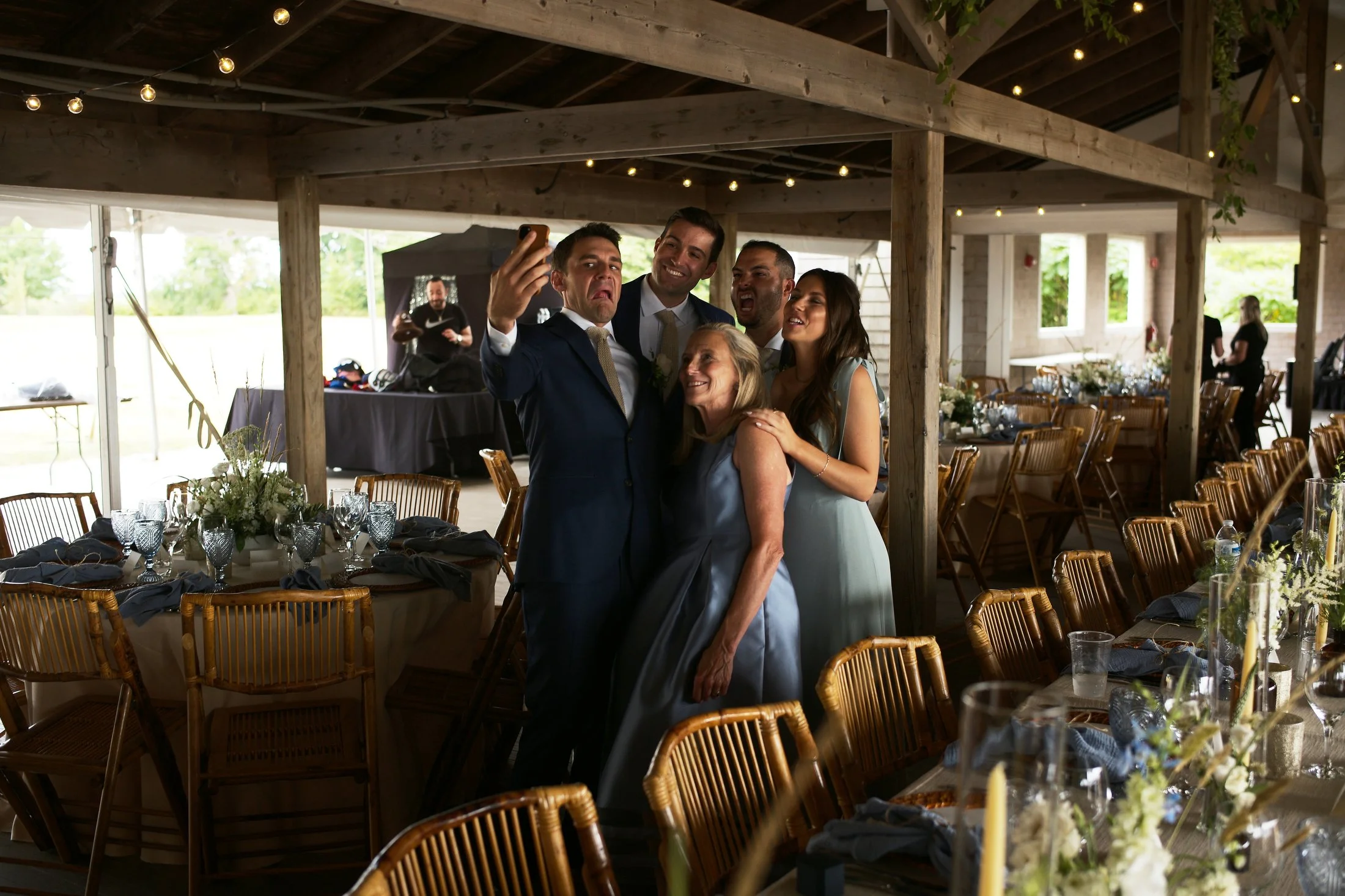 Group of five people taking a selfie at a wedding reception in a rustic decorated venue with tables, chairs, and floral centerpieces.