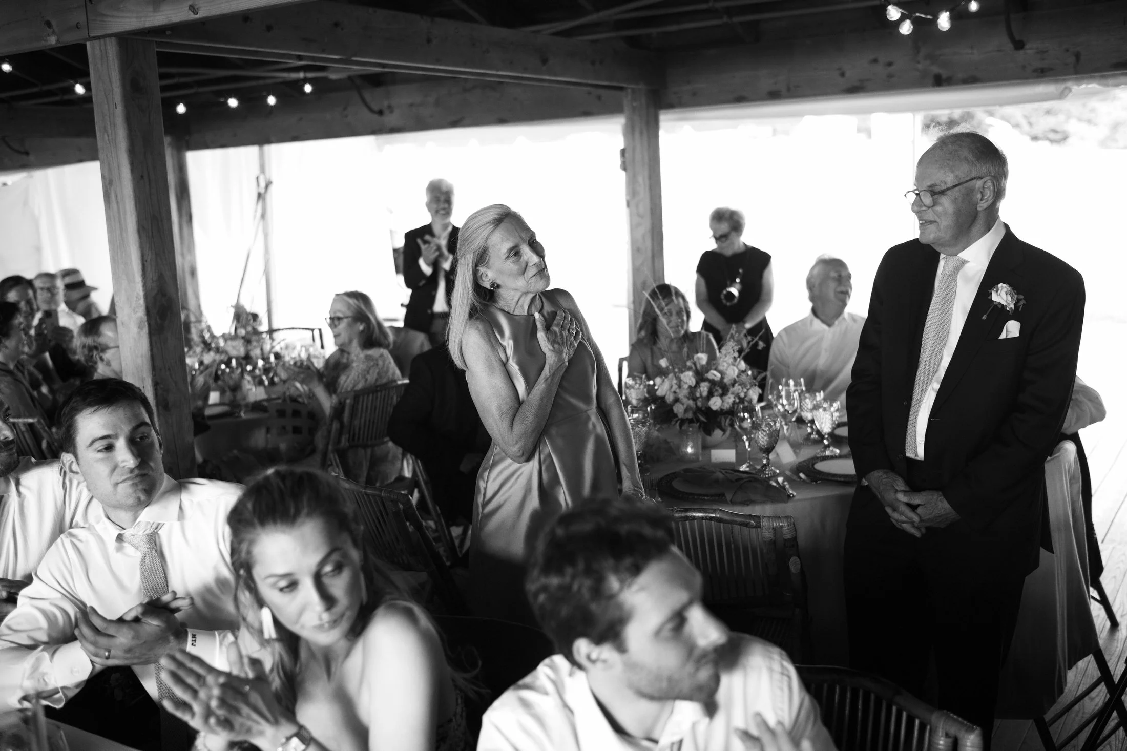 A black and white photo of a wedding reception with guests seated at tables. One woman stands, emotional, with her hand on her chest, facing a man in a tuxedo, who is smiling. Other guests are clapping and watching in the background, under a wooden c