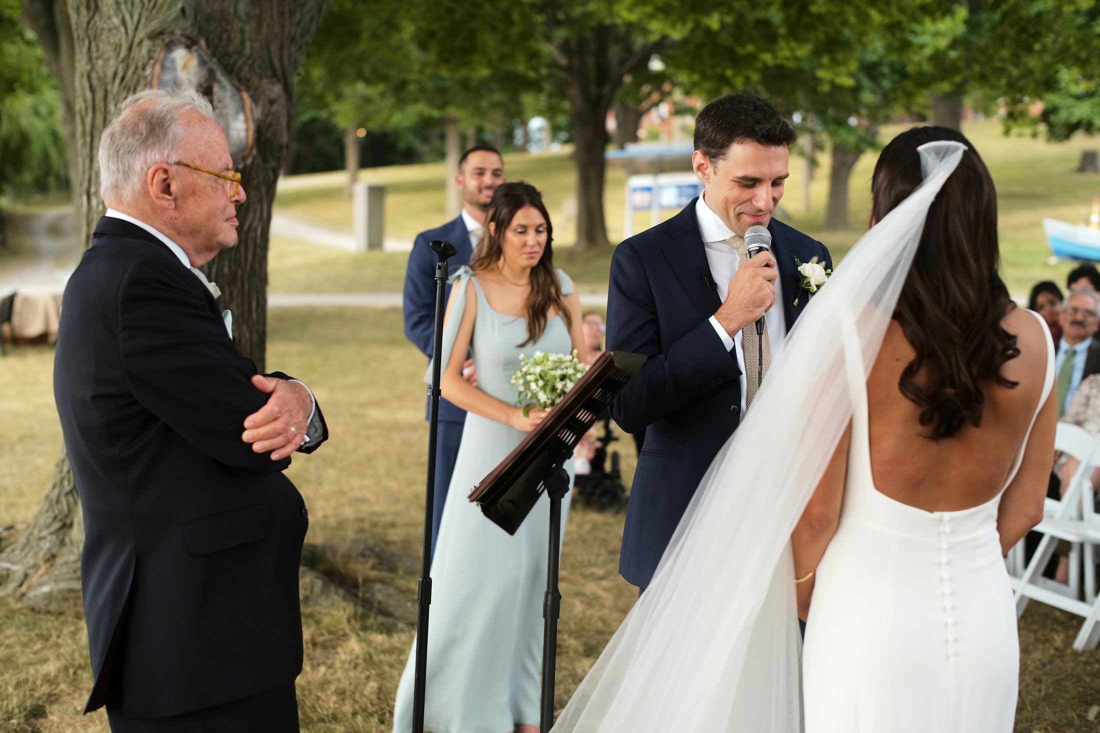 A wedding ceremony outdoors with a groom holding a microphone in front of the bride, who is wearing a white dress and veil. An older man in a tuxedo stands nearby with arms crossed. Bridesmaids and groomsmen, and seated guests are visible in the back