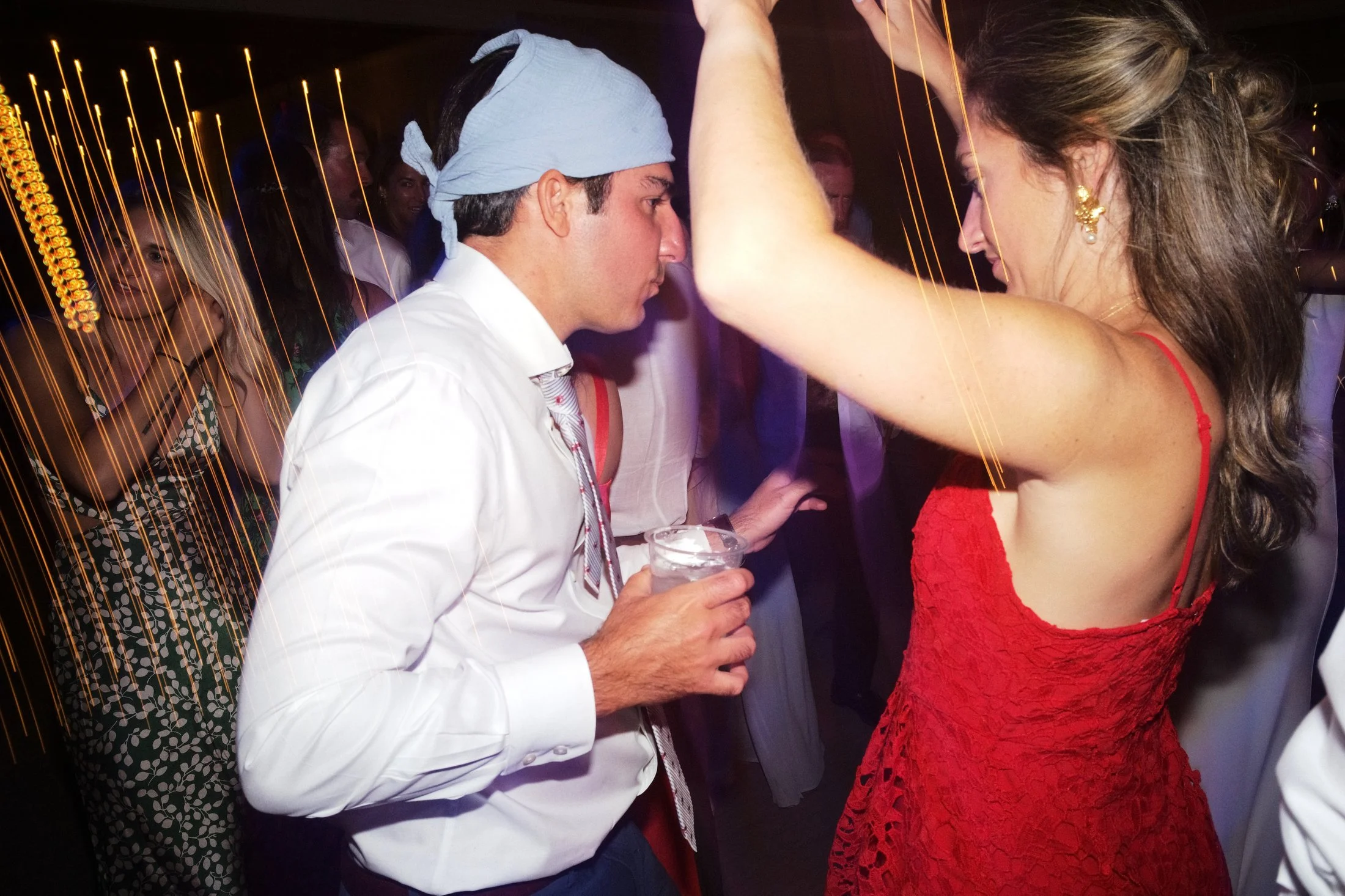A man in a white shirt and a light blue bandana on his head is holding a drink and is dancing closely to a woman in a red dress. The woman has long, wavy hair, gold earrings, and is raising her arm, looking down, while the man is leaning towards her 